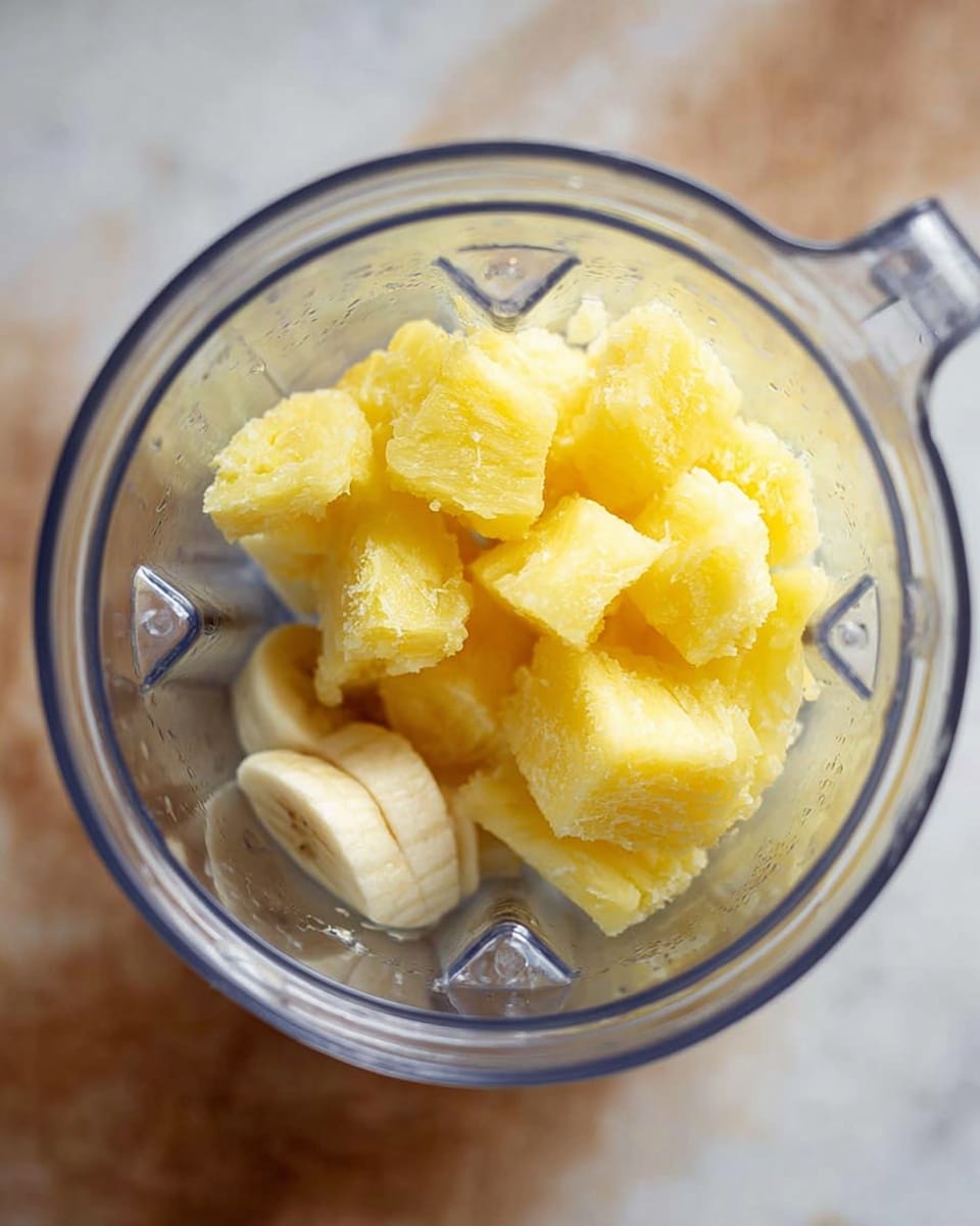 Inside a clear blender jar are two layers of ingredients. The bottom layer is soft and pale yellow, likely slices of banana with a smooth texture. On top, there is a thicker layer of bright yellow frozen pineapple chunks with a rough, icy texture. The blender jar is placed over a white marbled surface. Photo taken with an iphone --ar 4:5 --v 7