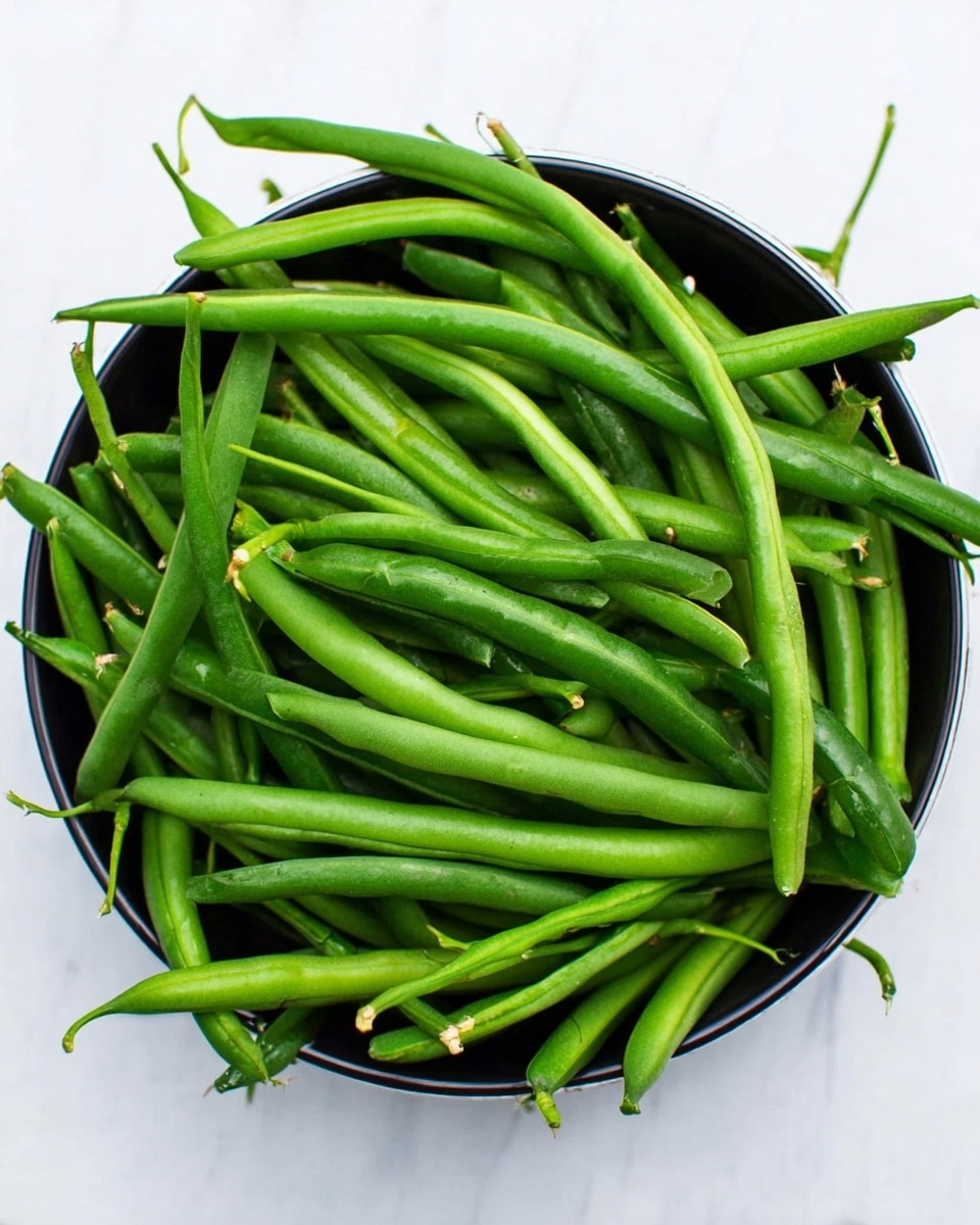A top view of a black bowl filled with many fresh green beans, the green beans are vibrant and smooth with some thin stems and tips visible. The bowl is placed on a white marbled surface, giving a clean and bright background to the fresh green beans. photo taken with an iphone --ar 4:5 --v 7