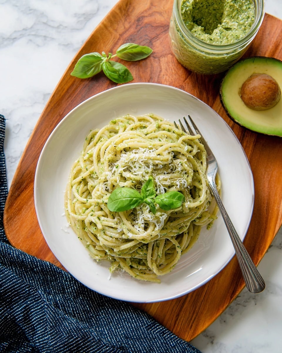 A white plate holds a single serving of creamy green pasta with visible flecks of herbs throughout, topped with bright green fresh basil leaves and some grated white cheese scattered gently on top. The plate sits on a warmly colored wooden board with a silver fork tucked in on the left side of the pasta. To the upper right, there is a jar filled with textured green sauce, and nearby sits half of an avocado with its light green inside and brown seed showing. The backdrop is a white marbled surface, and a dark blue striped cloth lies partially under the wooden board. photo taken with an iphone --ar 4:5 --v 7