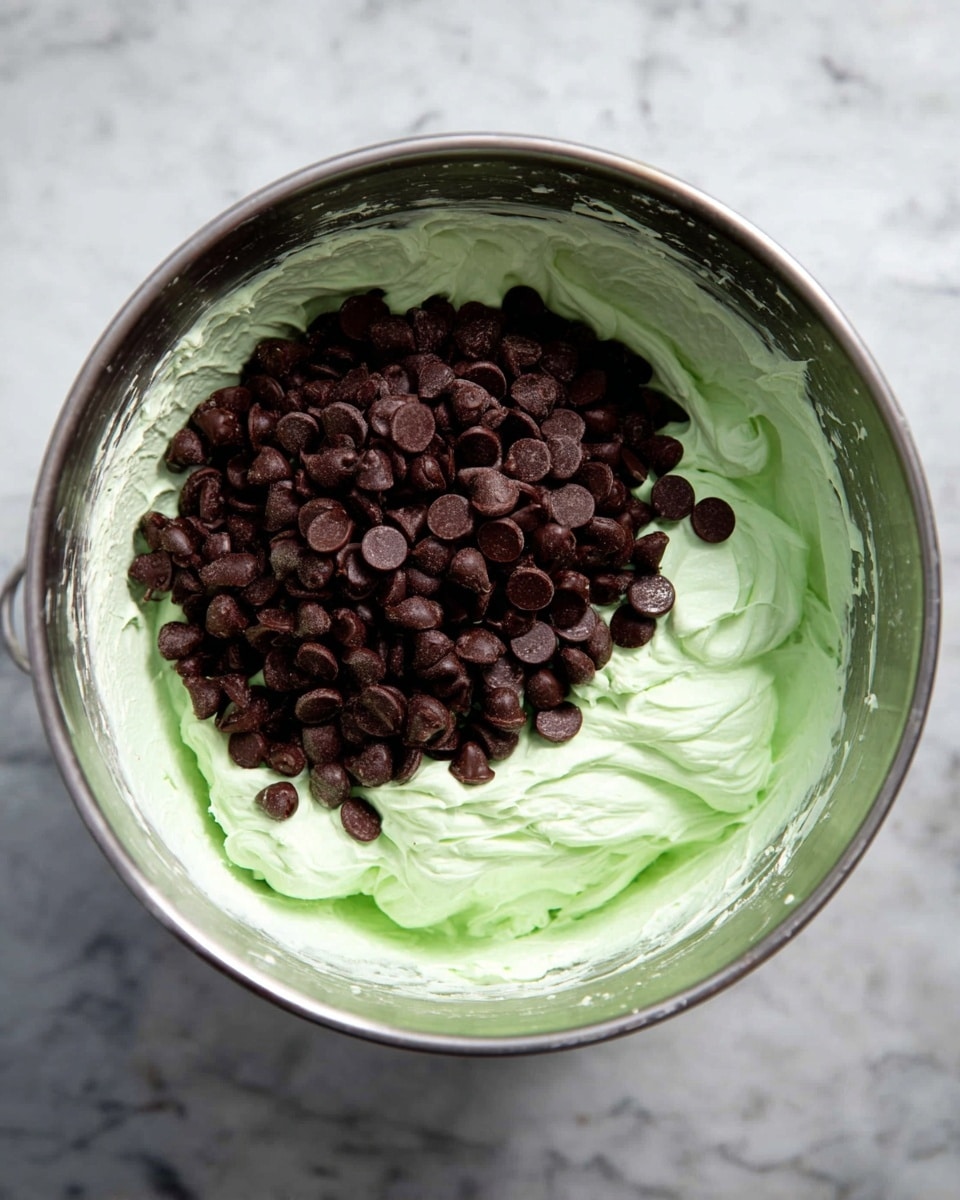A metal mixing bowl contains two main layers: the bottom layer is a light green whipped creamy mixture with a smooth, fluffy texture covering the bowl’s inner surface, and on top, a mound of dark brown chocolate chips is placed mostly in the center. The bowl is set on a white marbled surface, and the image is taken from directly above, showing the glossy shine inside the bowl. Photo taken with an iphone --ar 4:5 --v 7
