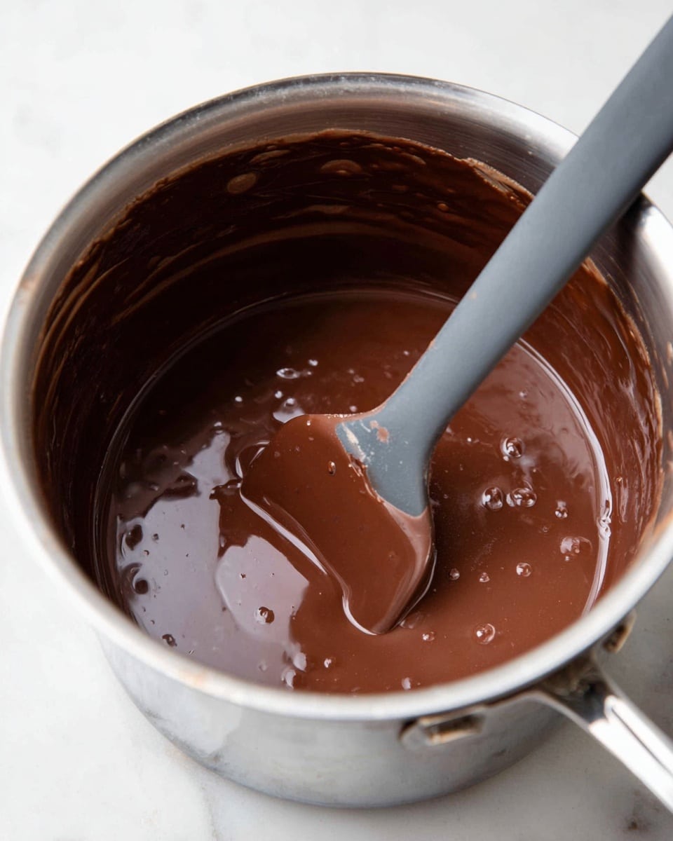 A close-up view of a shiny silver pot filled with smooth, glossy dark brown chocolate sauce that looks thick and creamy. Inside the pot is a gray silicone spatula partially covered in the rich chocolate, with the spatula's flat end dipped in the sauce, lifting some chocolate slightly above the surface. The pot sits on a white marbled surface, and small bubbles and streaks of chocolate line the inside walls of the pot, showing the sauce's silky texture. Photo taken with an iphone --ar 4:5 --v 7