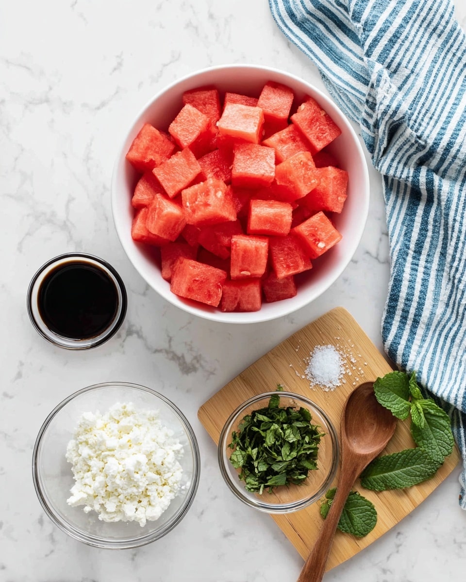 A white bowl filled with bright red watermelon cubes sits at the top left of the image on a white marbled surface. Below it are two small clear glass bowls, one containing dark balsamic vinegar and the other white crumbled cheese. To the right is a small wooden board holding a small clear bowl with chopped green herbs, a wooden spoon with coarse white salt, and a few fresh green mint leaves resting on the board. A blue and white striped cloth is draped in the top right corner, and everything is photographed clearly with natural light, photo taken with an iphone --ar 4:5 --v 7