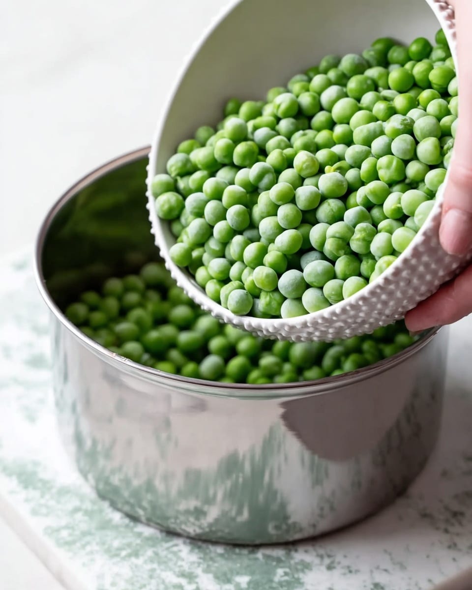 A close-up view showing a white bowl with textured edges filled with bright green frozen peas being tilted by a woman's hand over a shiny silver metal pot. The pot sits on a white marbled surface with green speckles. The scene is well-lit, highlighting the smooth and shiny metal of the pot and the round shapes of the frozen peas. photo taken with an iphone --ar 4:5 --v 7
