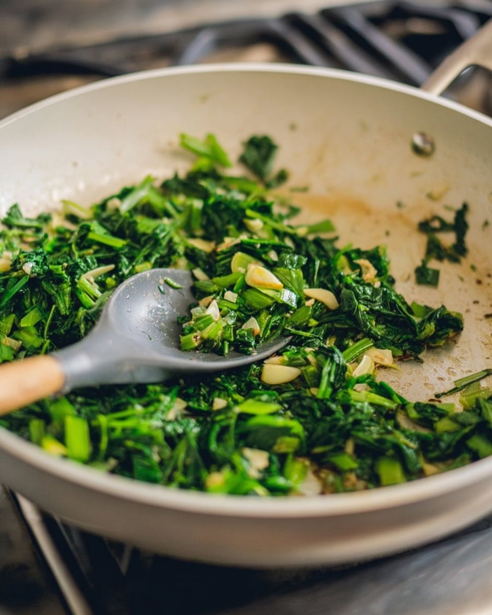 A white frying pan with a mix of chopped green leafy vegetables and small pieces of garlic cooking inside. The greens are bright and fresh, with some pieces slightly softened and others still firm. A gray spoon with a light wooden handle rests inside the pan, partially touching the greens. The pan is on a stove with a blurred background, and the cooking surface beneath appears stained and worn. photo taken with an iphone --ar 4:5 --v 7
