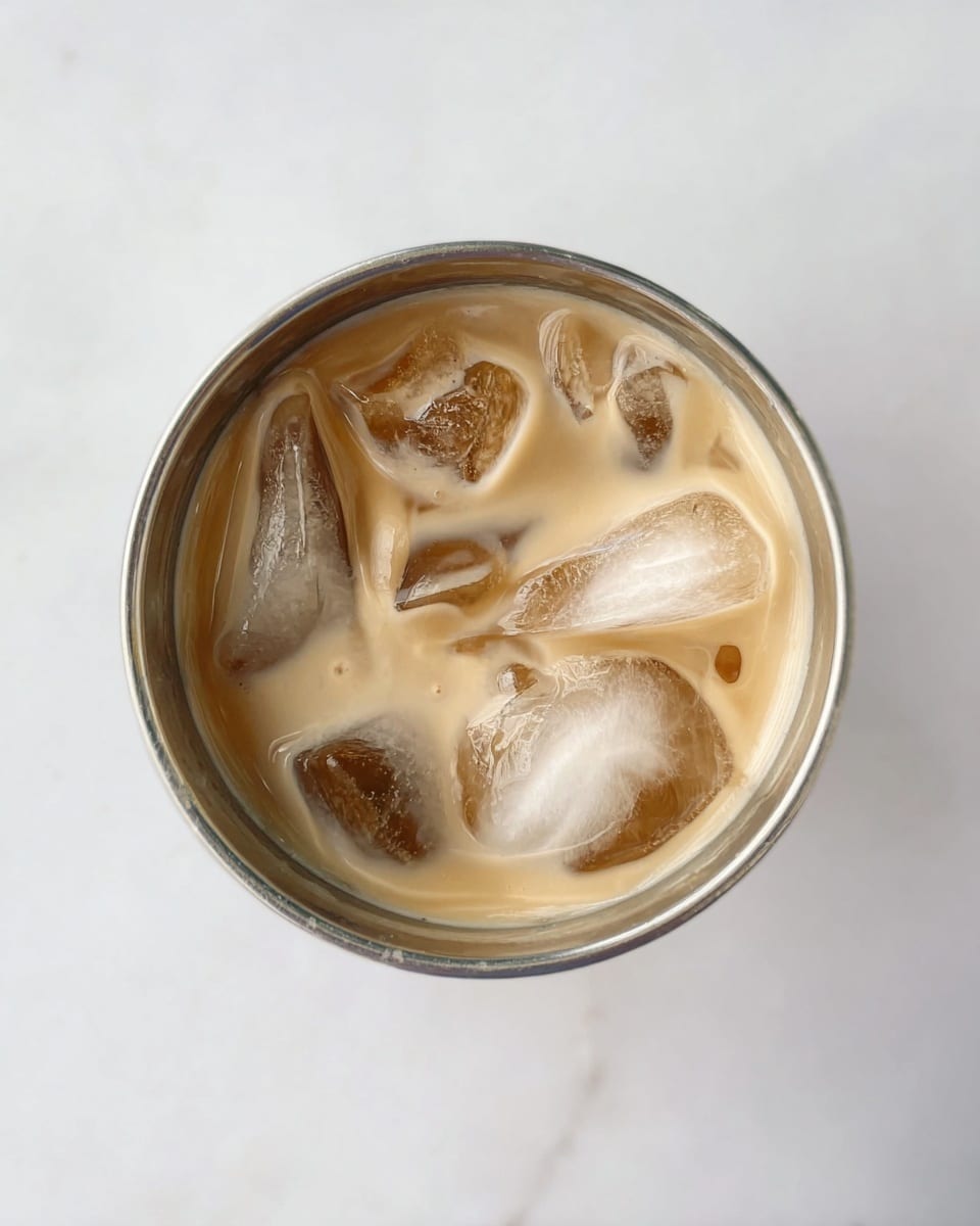A metal cup is filled with a creamy light brown drink and several chunky ice cubes floating on the top. The ice cubes are clear with some white cloudy parts inside, and they sit randomly in the liquid. The cup is centered on a white marbled surface that makes the light drink color stand out. photo taken with an iphone --ar 4:5 --v 7