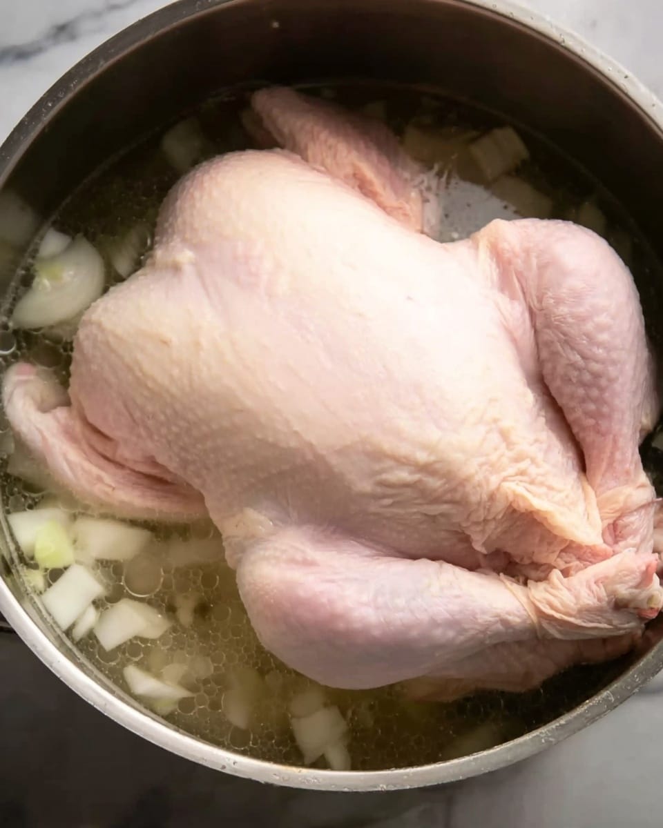 A whole raw chicken with pale pink skin is placed inside a large metal pot filled with clear liquid, with chopped white pieces, likely onions, scattered nearby in the broth. The chicken is centered and fully submerged, with visible wings and legs slightly lifted above the liquid. The background shows the inside of the metal pot and a white marbled surface under it. Photo taken with an iphone --ar 4:5 --v 7