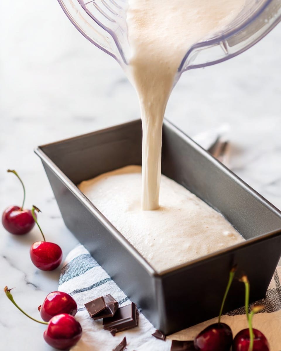 A light cream-colored batter is being poured from a clear blender into a rectangular grey metal loaf pan. The batter has a smooth, slightly frothy texture as it flows down into the pan, filling it evenly. Around the pan on the white marbled surface are several bright red cherries with green stems and small pieces of dark brown chocolate. A white and blue striped cloth is partially visible under the pan and cherries. The setting is bright and clean, with soft natural light highlighting the textures and colors photo taken with an iphone --ar 4:5 --v 7