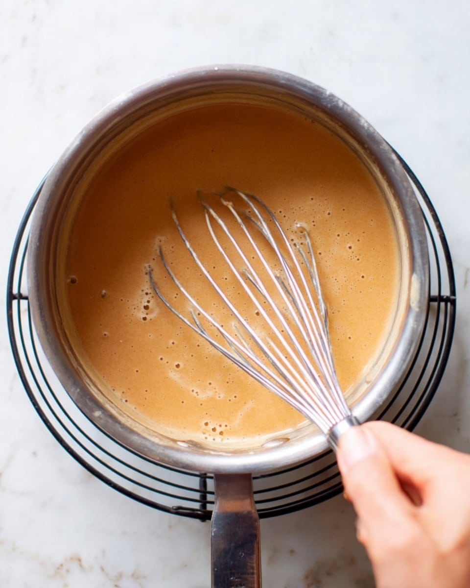 A close-up of a silver saucepan filled with a smooth, light brown and creamy sauce being gently stirred with a metal whisk held by a woman's hand. The saucepan is placed on a black wire cooling rack, which sits on a white marbled surface. The sauce has a slight shine and small bubbles, showing it is being heated and mixed evenly. The woman's hand is positioned on the whisk handle stirring the sauce inside the pan. photo taken with an iphone --ar 4:5 --v 7