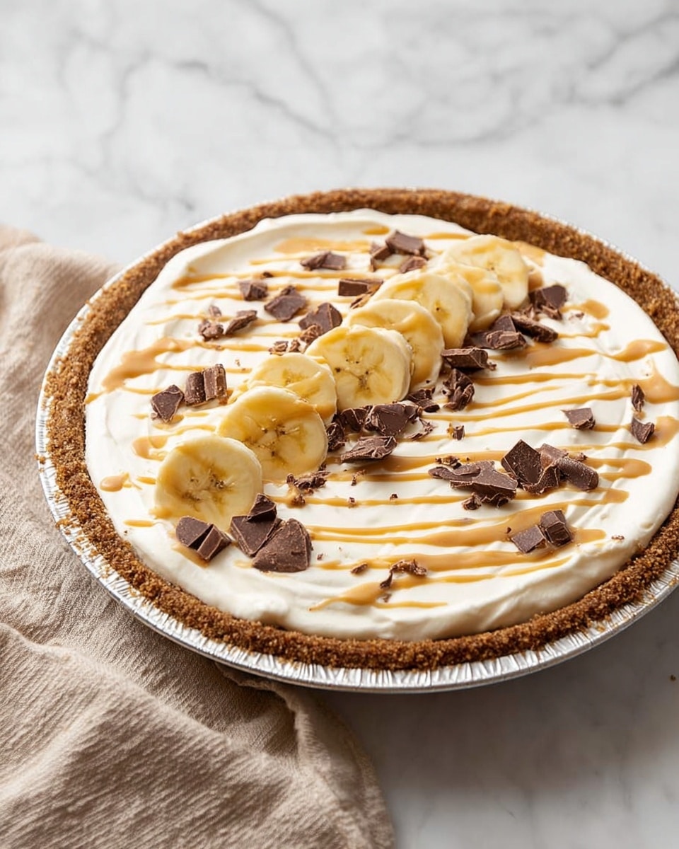 The image shows a round pie with three main layers. The bottom layer is a thick, brown crust that looks crumbly and firm, forming the base and edges of the pie. On top of this is a thick, smooth white cream layer that spreads evenly across the pie. In the center, there is a line of banana slices placed diagonally from the bottom left to the top right, bright yellow with a soft texture. Scattered around and on top of the cream are small dark brown chocolate chunks. Over everything, a light brown sauce is drizzled in thin, uneven lines from left to right. The pie is in a round metal pan sitting on a white marbled surface with a beige cloth beside it. photo taken with an iphone --ar 4:5 --v 7