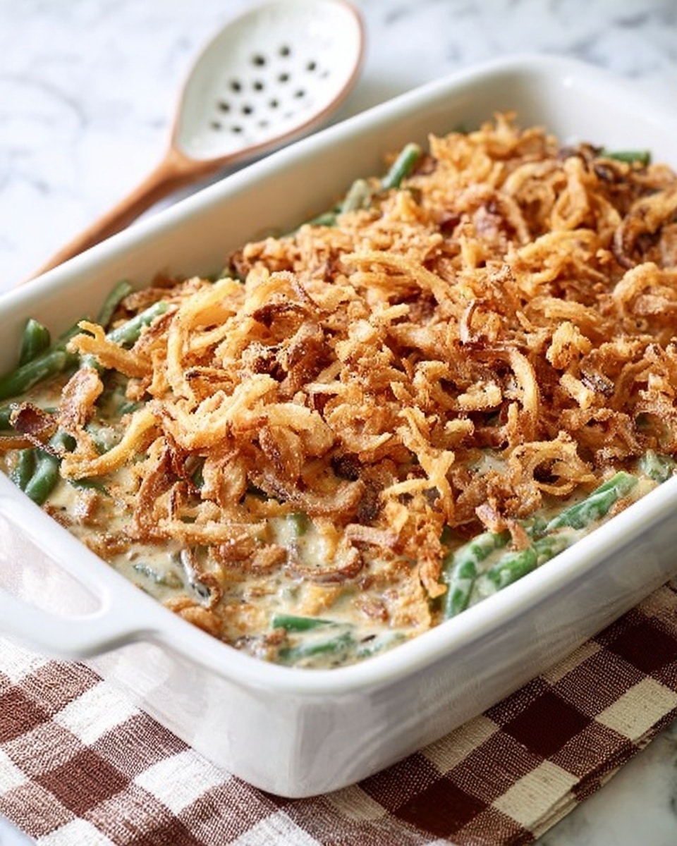 A rectangular white dish filled with a creamy green bean casserole topped with a thick, even layer of golden brown crispy fried onions. The green beans and creamy sauce mixture underneath the topping peek through slightly at the edges, showing soft green and beige hues. The dish sits on a checkered cloth with a white slotted spoon nearby, all set against a white marbled surface. Photo taken with an iphone --ar 4:5 --v 7