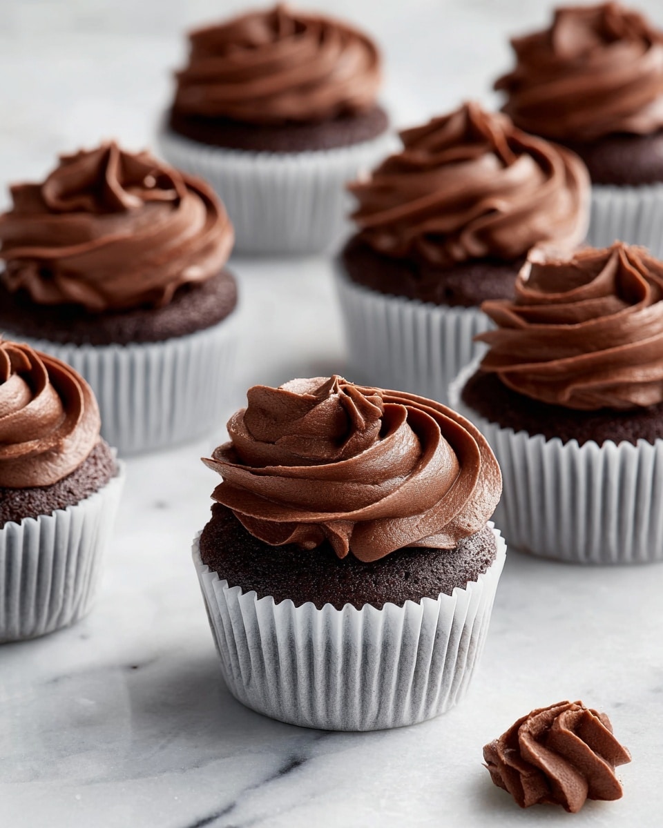 The image shows several chocolate cupcakes placed on a white marbled surface. Each cupcake has two layers: a dark brown chocolate base wrapped in a white paper liner, and a thick swirl of smooth, creamy chocolate frosting on top. The frosting has a rich, glossy texture with soft peaks and curves, forming a rose-like pattern. The cupcakes are arranged so that some are close while others are blurred further away, adding depth to the scene. In the bottom right corner, a small dollop of chocolate frosting sits on the surface. Photo taken with an iphone --ar 4:5 --v 7
