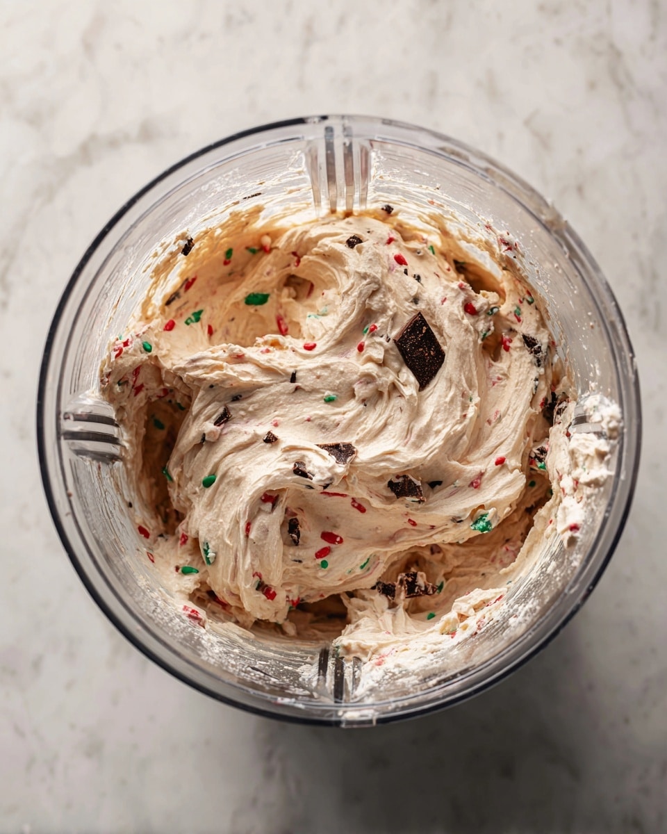 Inside a clear blender jar, there is a creamy mixture with a light beige color. Mixed throughout the creamy base are small pieces of dark chocolate and bits of red and green candy, adding bright touches of color. The texture of the mixture looks thick and slightly soft, with some swirls and folds showing its whipped nature. The blender jar has vertical ridges and a rounded shape, sitting on a white marbled surface. photo taken with an iphone --ar 4:5 --v 7
