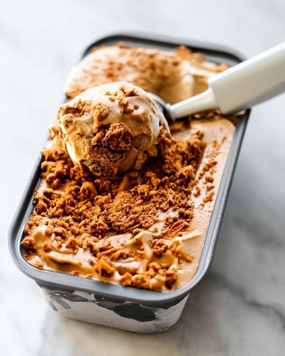 A close-up view of a metal container filled with a smooth, light brown ice cream base topped with a generous layer of crumbly, darker brown crunchy pieces. A single round scoop of the ice cream with crumbly bits attached is lifted by a white ice cream scooper, resting on top of the layer. The texture of the ice cream looks creamy with rough crunchy bits scattered across the surface. The container sits on a white marbled surface with a bright, clean background, giving a fresh look. Photo taken with an iphone --ar 4:5 --v 7