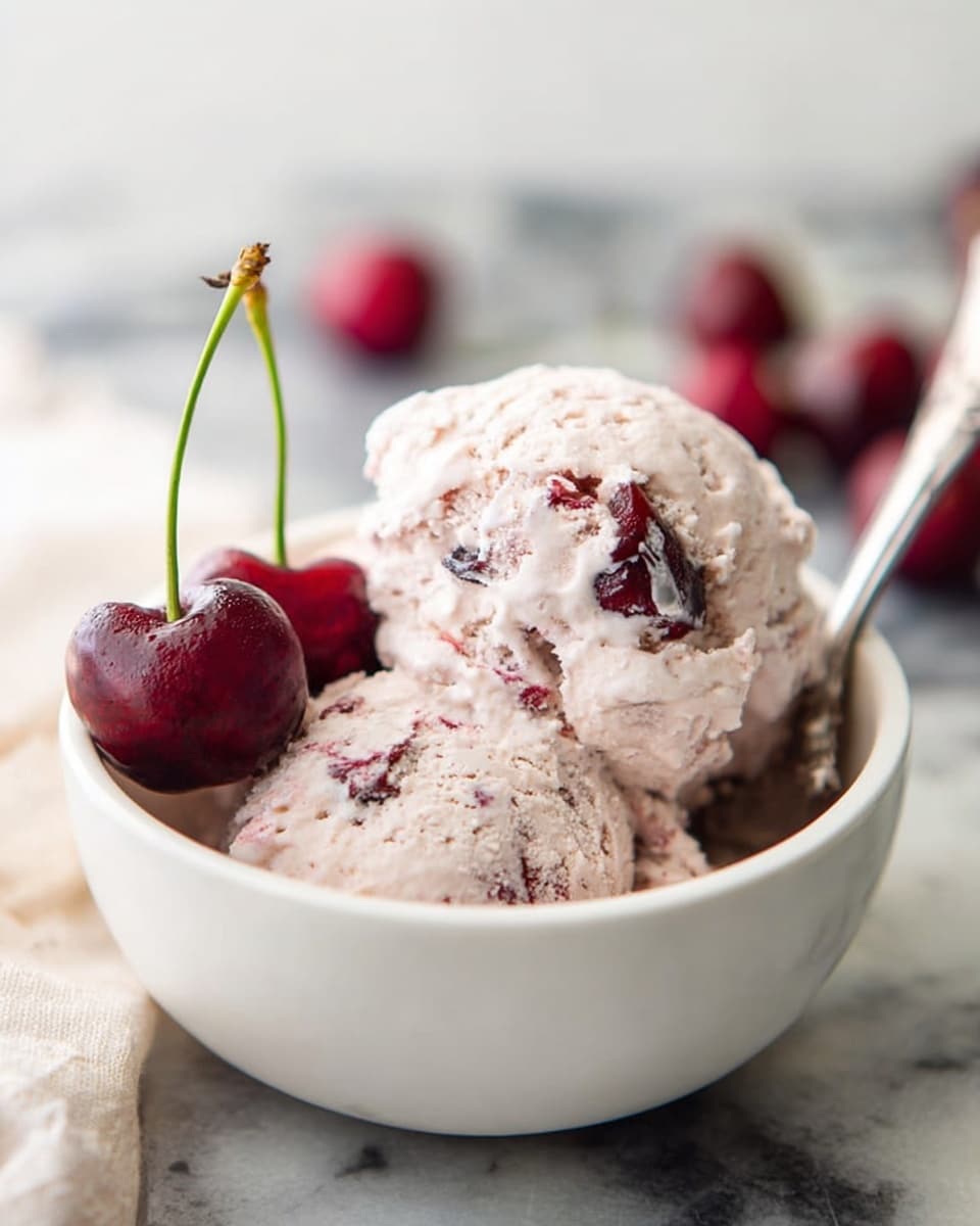 A white bowl holds two scoops of pale pink ice cream with visible pieces of dark red cherries mixed inside. On the left side of the bowl, two fresh, whole cherries with green stems rest against the ice cream. A silver spoon lies inside the bowl on the right side, slightly under the ice cream. The bowl sits on a white marbled surface with a soft, blurred background where some more cherries can be seen out of focus. Photo taken with an iphone --ar 4:5 --v 7