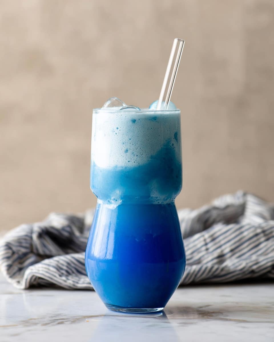 A vibrant blue creamy drink fills a three-layered clear glass with a wide middle and narrow base and top, showing visible ice cubes inside. The top layer has a frothy light blue foam that softly overlaps the glass's edge. A transparent straw stands in the drink extending upward. The glass sits on a white marbled surface with a soft beige blurred background, and there is a striped cloth partially visible in the back. photo taken with an iphone --ar 4:5 --v 7