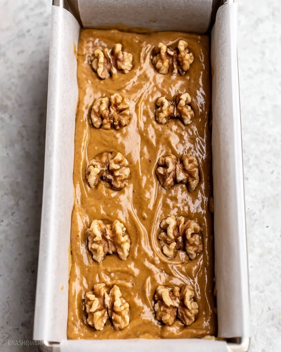 The image shows a rectangular white loaf pan filled with a thick, light brown batter that looks smooth and creamy. On top of the batter, there are evenly spaced walnut halves, ten in total, arranged in two rows of five, with a rough texture contrasting the smooth batter. The pan is lined with white parchment paper sticking out on the sides. The pan rests on a white marbled textured surface, with soft natural light highlighting the glossy detail of the batter and the rough texture of the walnut pieces. Photo taken with an iphone --ar 4:5 --v 7