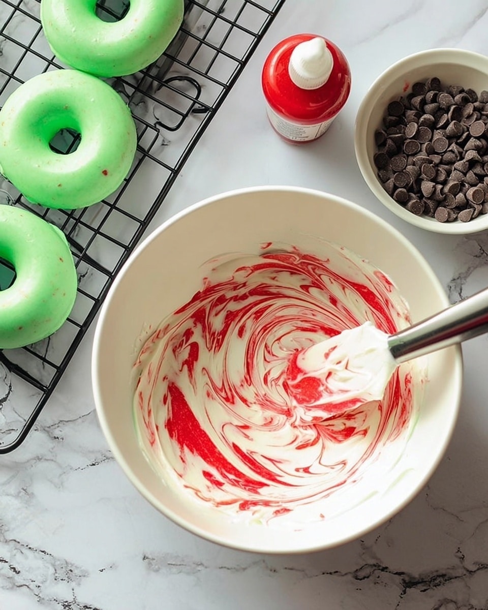 The image shows a white bowl with white creamy frosting mixed with bright red syrup swirled on the top, creating a marbled red and white pattern. A spatula with some frosting is placed inside the bowl, and next to it, on a white marbled surface, there is a small bottle of red food coloring and a white bowl filled with small dark chocolate chips. To the left, two green frosted round donuts with holes in the center are resting on a black cooling rack. Photo taken with an iphone --ar 4:5 --v 7