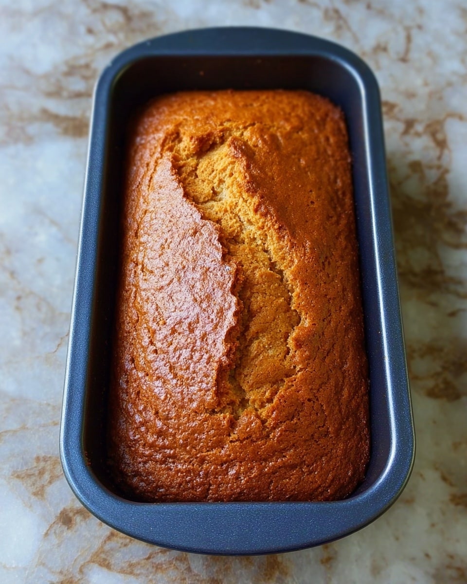 A golden-brown loaf of bread with a cracked top sits inside a dark blue baking tin, showing a slightly rough texture on the crust. The loaf fills the tin completely and looks soft yet firm. The baking tin is placed on a white marbled surface with a subtle pattern, highlighting the warm, baked colors of the bread. photo taken with an iphone --ar 4:5 --v 7