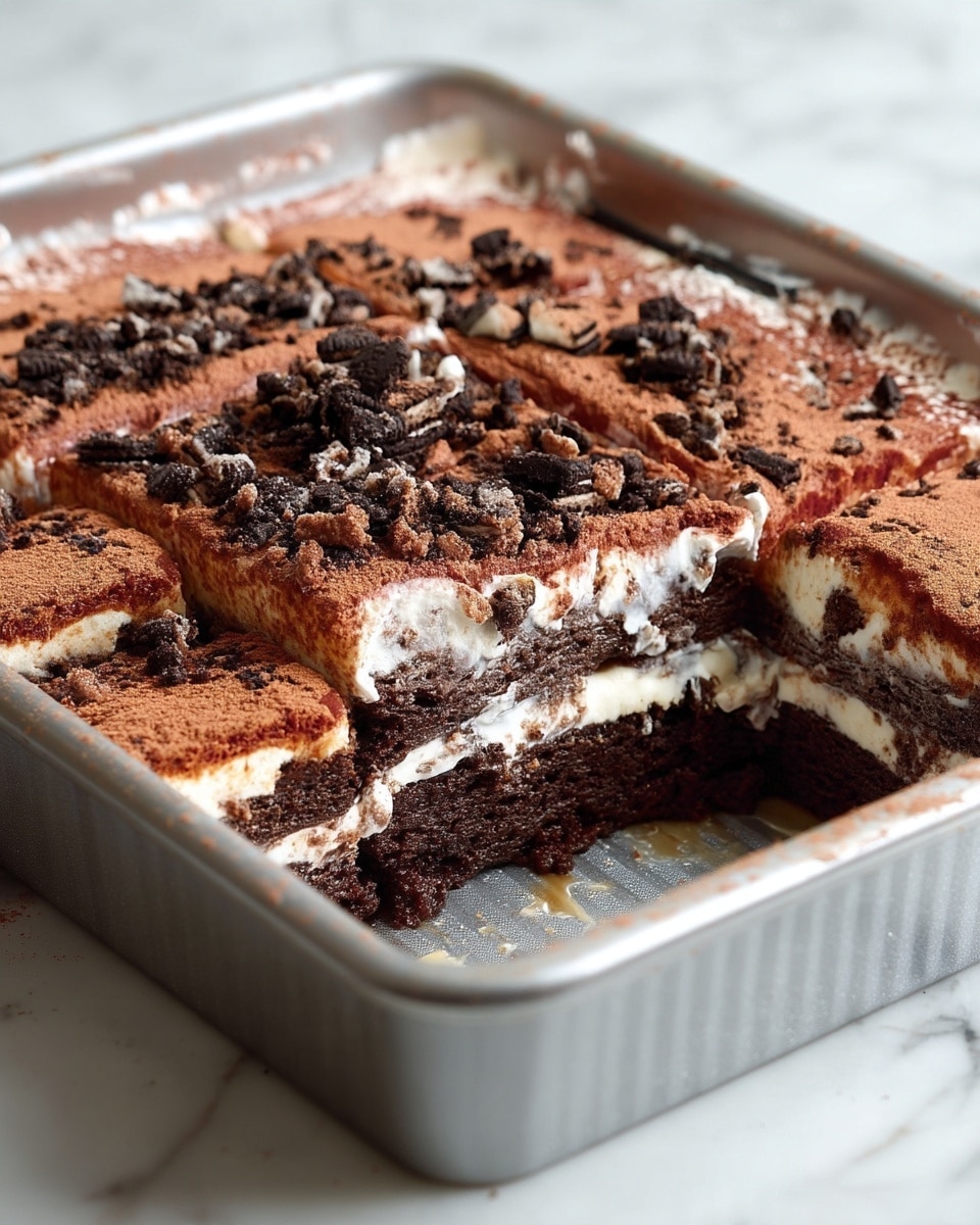 The image shows a close-up of a square chocolate dessert in a metal baking tray with one portion removed, revealing three layers: a bottom thick, moist dark brown cake layer, a middle white creamy layer with small bits of dark crumbs mixed in, and a top thick creamy layer dusted with a light brown cocoa powder and sprinkled with dark crushed cookie pieces. The dessert is cut into nine squares, and some crumbs and syrup spots are visible inside the empty space where one piece was taken out. The scene has a white marbled surface in the background. photo taken with an iphone --ar 4:5 --v 7