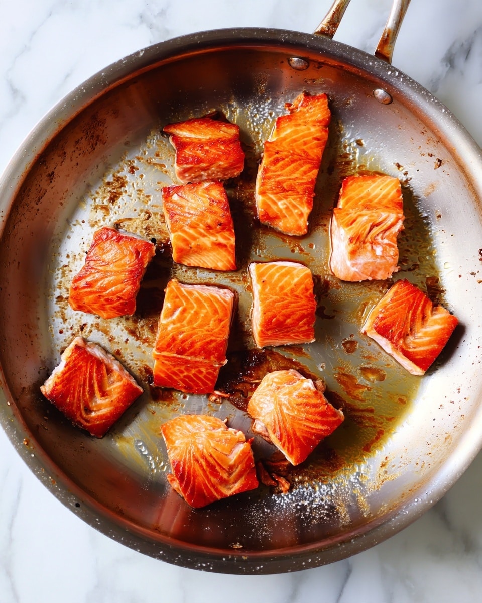 Pieces of cooked salmon are spread out in a shiny silver pan with some browned bits and slight oil shininess on the pan's surface. The salmon pieces are medium-sized, with a rich orange color and crisp, slightly charred edges showing a texture of cooked fish layers. The pan is on a white marbled background. photo taken with an iphone --ar 4:5 --v 7