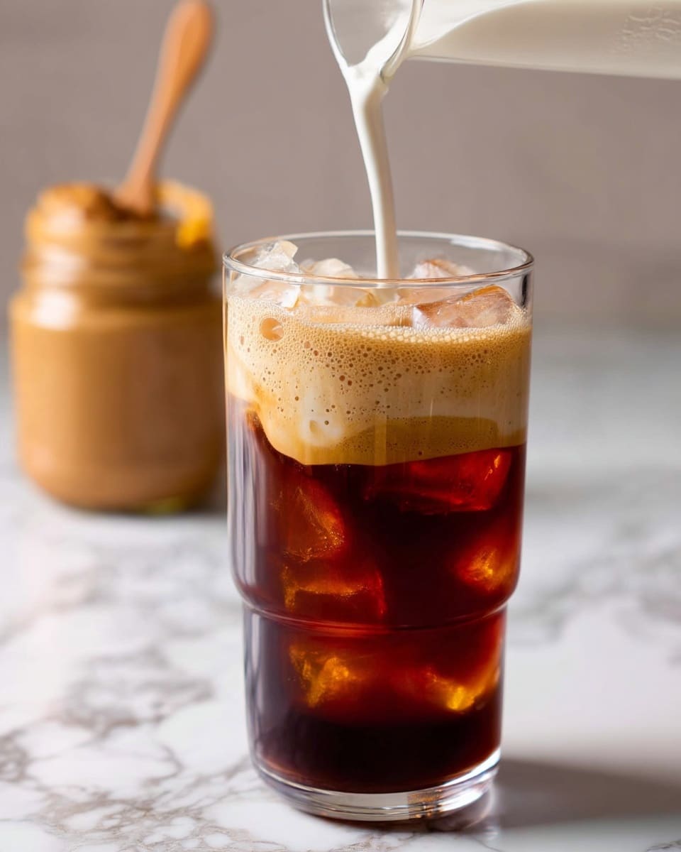 A clear glass with three layers stands on a white marbled surface: the bottom layer is dark brown cold coffee with ice cubes visible inside, the middle layer is a light foam of coffee crema, and the top layer is white creamy milk being poured from above, creating soft ripples on the surface. In the blurred background, a jar with a wooden spoon and some brown cream inside can be seen, adding a cozy kitchen feel. The photo taken with an iphone --ar 4:5 --v 7