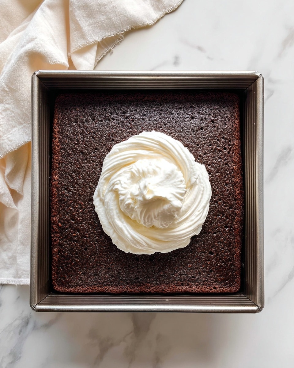 The image shows a square metal baking pan with a single layer of dark brown chocolate cake inside. In the center of this cake layer, there is a dollop of white whipped cream with a soft, fluffy texture. The pan sits on a white marbled surface, and a cream-colored cloth is partially visible on the upper left side. photo taken with an iphone --ar 4:5 --v 7