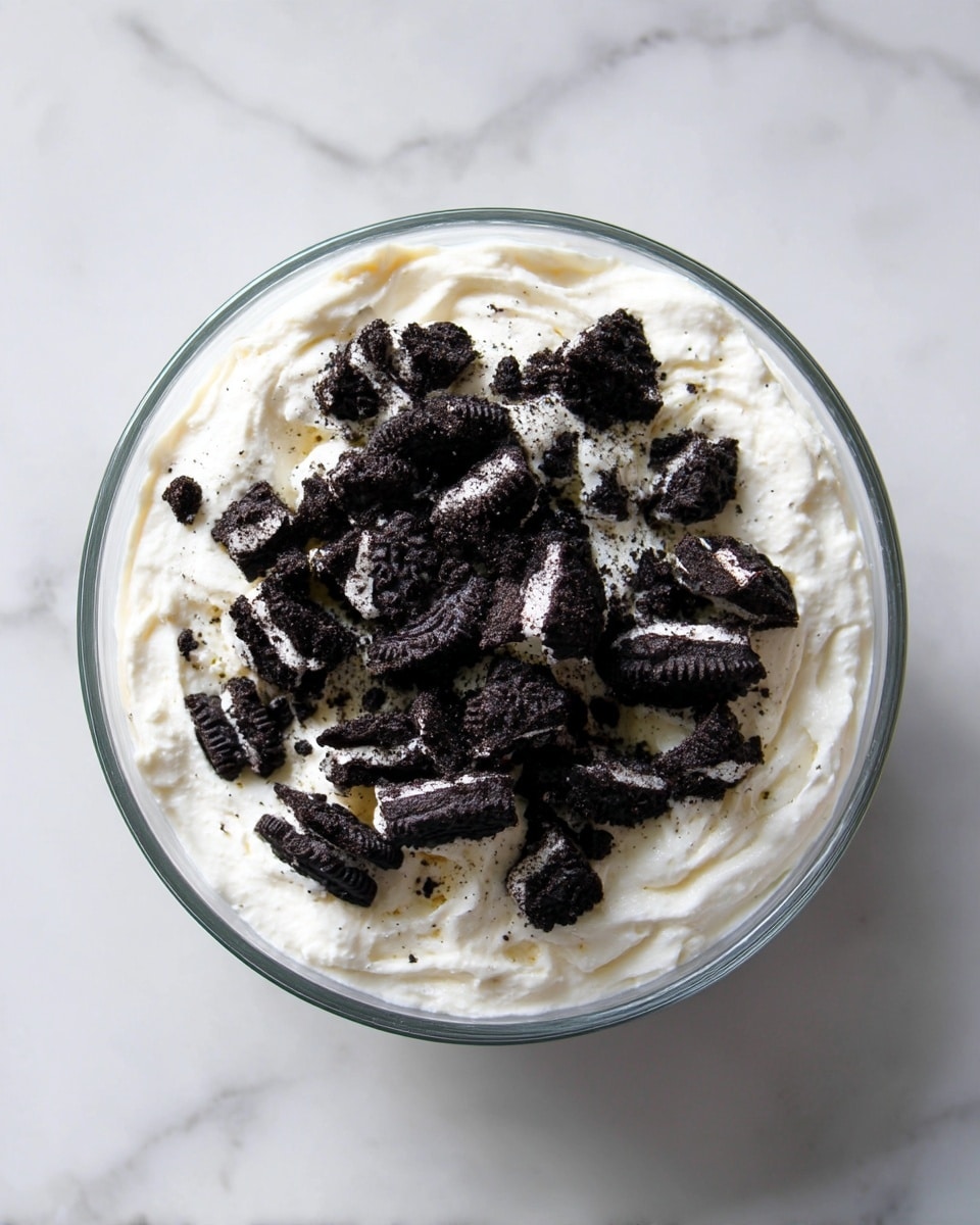 A clear glass bowl filled with two main layers: the bottom layer is white cream with a soft, fluffy texture, and the top layer is made up of broken pieces of dark black chocolate cookies with white cream filling inside, scattered unevenly over the cream. The bowl sits on a white marbled surface, and the image is taken from above showing the contrast between the dark cookie pieces and the bright white cream. photo taken with an iphone --ar 4:5 --v 7