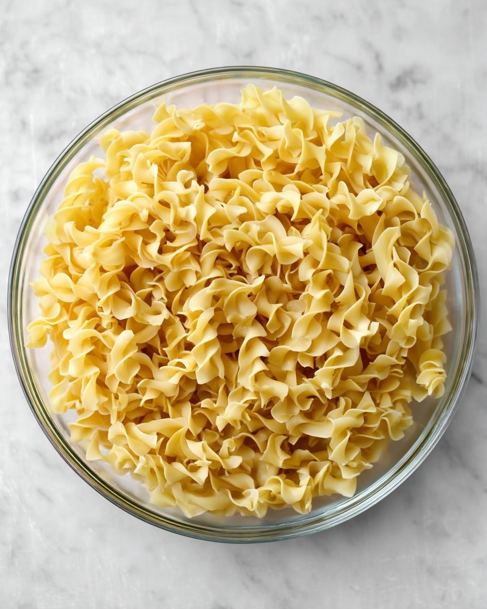 A clear glass bowl filled with twisted yellow egg noodles, showing their soft, curly texture in one even layer. The bowl sits on a white marbled surface with gentle natural light highlighting the smooth, slightly glossy noodles, arranged loosely but full to the top edges of the bowl, with the bowl’s transparent sides revealing the depth of noodles inside photo taken with an iphone --ar 4:5 --v 7