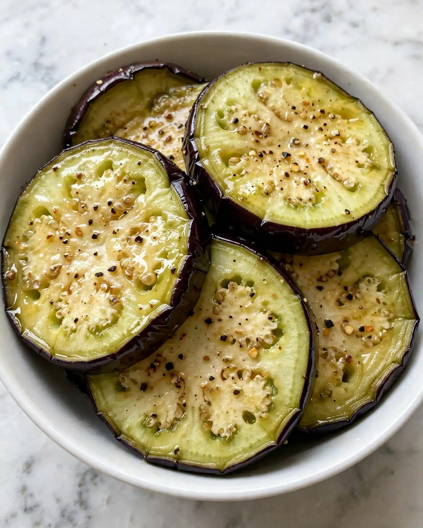 The image shows several thick, round slices of eggplant arranged in a white bowl. Each slice has a greenish-yellow center with a slightly spongy texture and dark purple edges from the skin. The slices are sprinkled with small black and brown pepper flakes and appear to have a light coating of oil, giving them a shiny look. The eggplants overlap each other slightly, filling most of the bowl. The bowl sits on a white marbled surface. Photo taken with an iphone --ar 4:5 --v 7