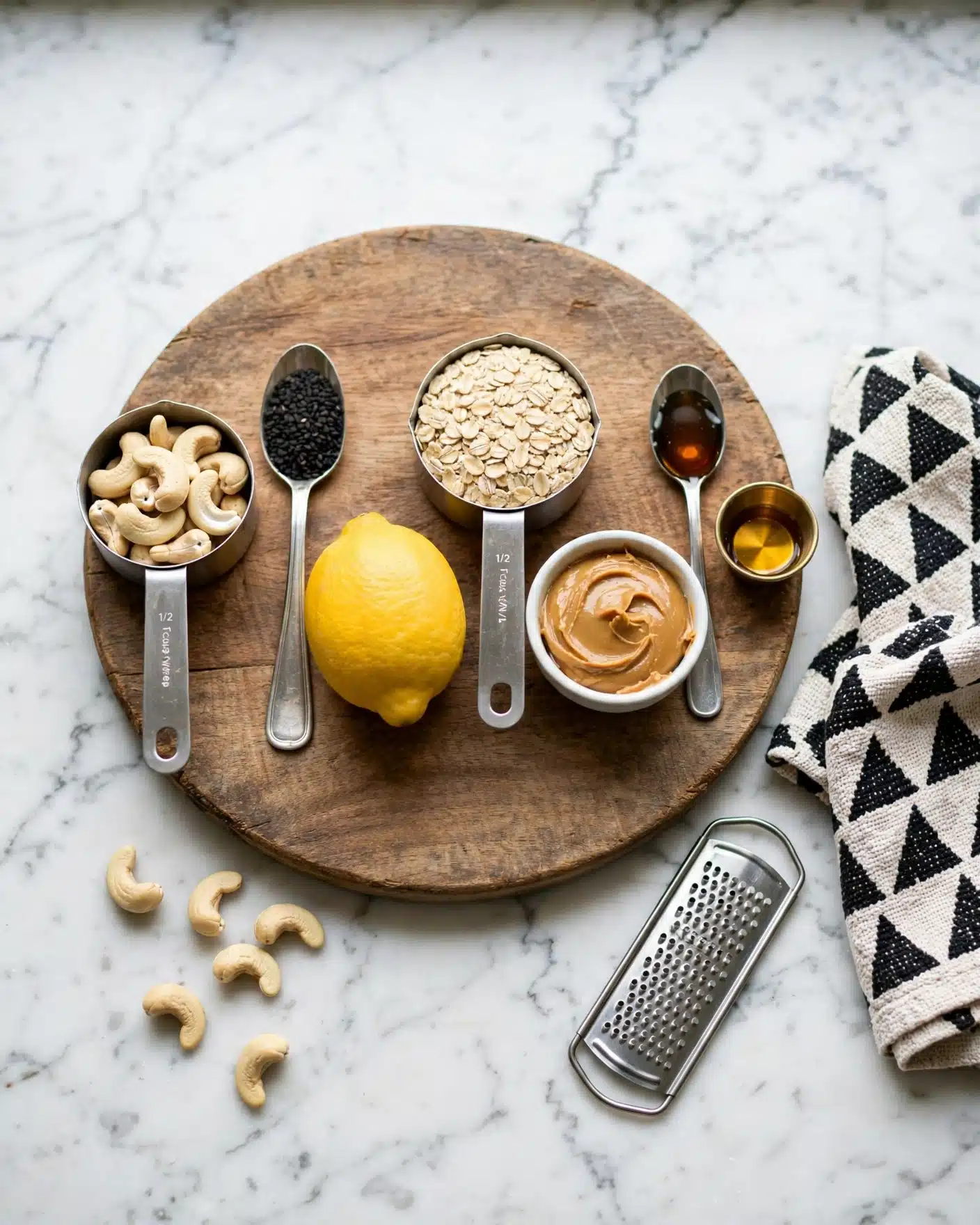 Lemon Poppyseed Energy Bites Recipe 4 A wooden round board sits on a white marbled surface, holding six measuring spoons and cups with different ingredients. From left to right: a ½ cup measuring cup filled with pale cashew nuts, a 1 tablespoon silver spoon filled with small black seeds, a whole bright yellow lemon in the center, a 1 cup measuring cup full of light tan rolled oats, a white small bowl filled with smooth light brown peanut butter, a 1 teaspoon silver spoon holding dark amber liquid, and a tiny metal cup with a small amount of golden liquid. A silver grater and a woven cloth with black triangles are nearby. A few cashew nuts lie on the white marbled surface near the board. Photo taken with an iphone --ar 4:5 --v 7