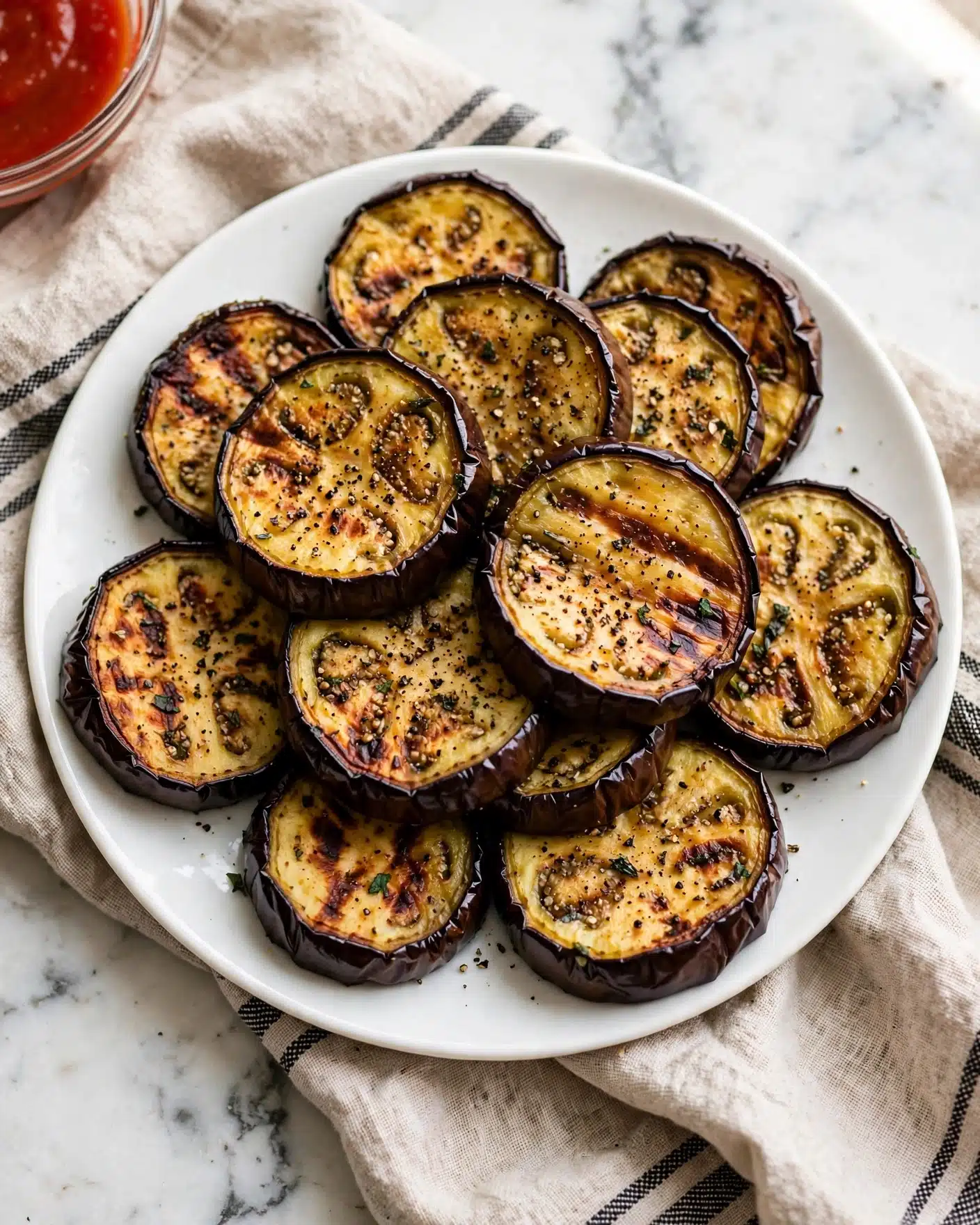 A white round plate holds about eleven thick slices of roasted eggplant arranged in a loose pile. Each slice has a dark purple, slightly wrinkled edge with a soft, golden-yellow center that shows some light browning and small grill marks. The surface of the eggplant slices is sprinkled with coarse black pepper and small green herb pieces for garnish. The plate sits on a beige linen cloth with black stripes, all placed on a white marbled surface. A small clear bowl with bright red sauce is partially visible at the top left corner of the image. photo taken with an iphone --ar 4:5 --v 7
