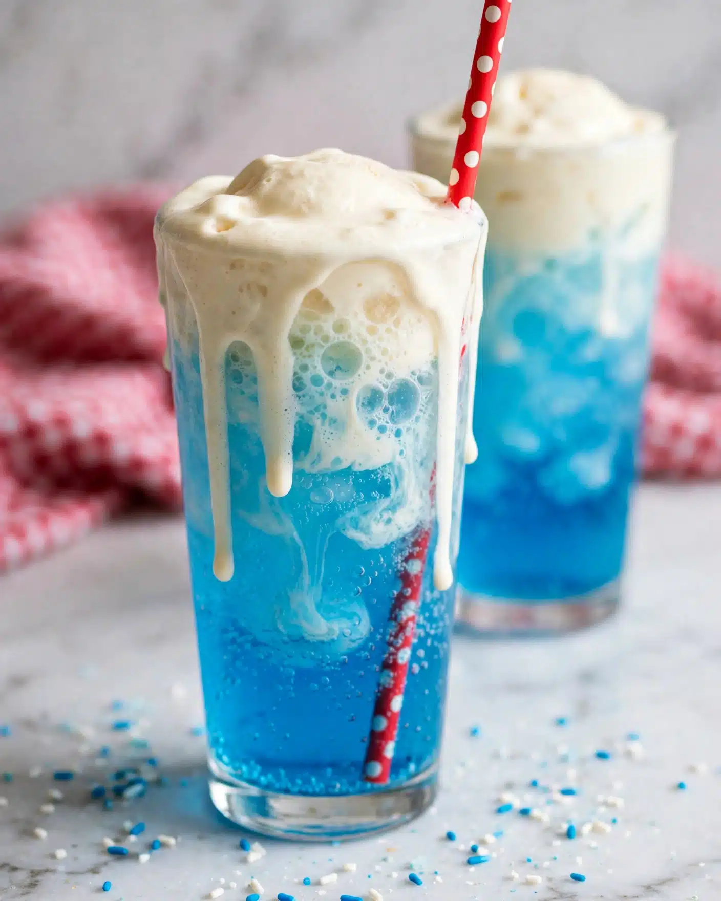 A clear glass holds a bright blue fizzy drink with a thick layer of white ice cream floating on top, creating a creamy white foam that blends down into the blue liquid. A red straw with white polka dots stands inside the glass on the right side, piercing the ice cream. In the background, another similar glass is softly focused on a white marbled surface with a red cloth slightly blurred behind. The drink looks cold and refreshing with bubbly texture near the top. photo taken with an iphone --ar 4:5 --v 7