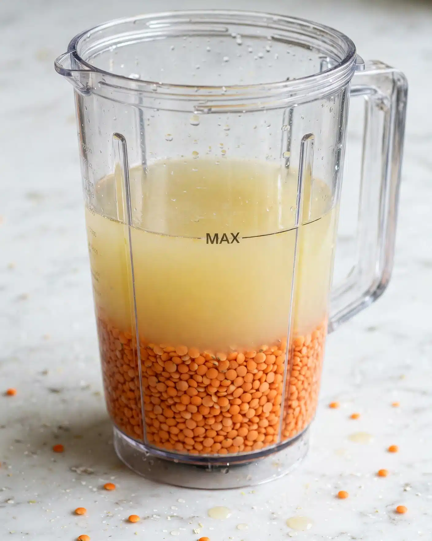 The image shows a clear blender container filled with two layers. The bottom layer consists of small, round orange lentils packed closely together. The top layer is a light, pale yellowish liquid that fills the container up to a 