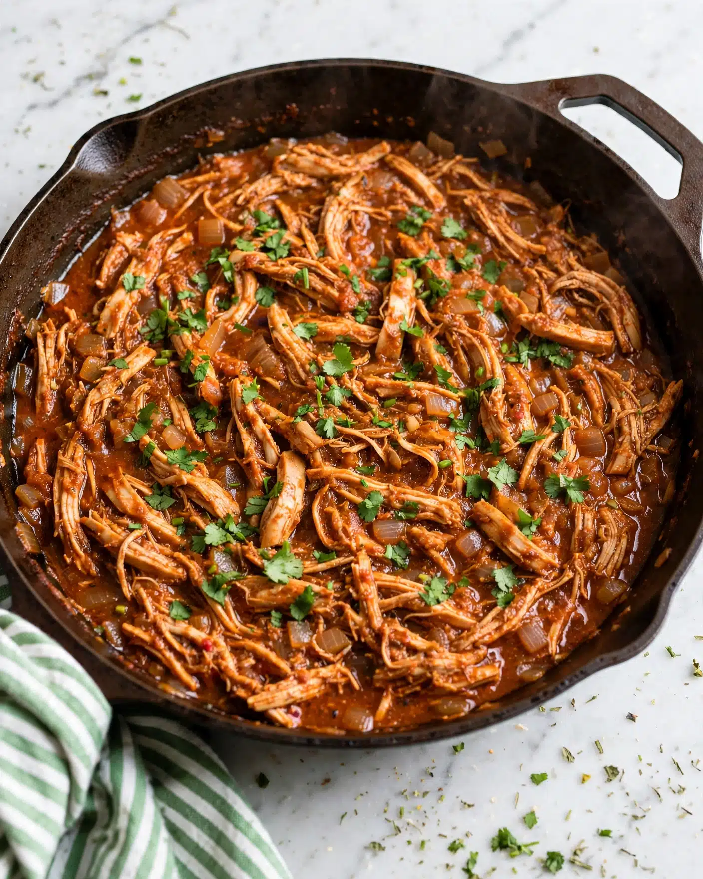 Keto Chicken Enchiladas Bowl Recipe 4 A large brown skillet is filled with shredded chicken mixed with a thick, rich reddish-brown sauce. The chicken is spread evenly in the pan with visible soft, fibrous texture and small bits of cooked onions throughout. Bright green chopped herbs are sprinkled on top, adding a fresh contrast to the warm colors of the dish. The skillet rests on a white marbled surface with a green and white striped cloth partially visible under one side. Photo taken with an iphone --ar 4:5 --v 7