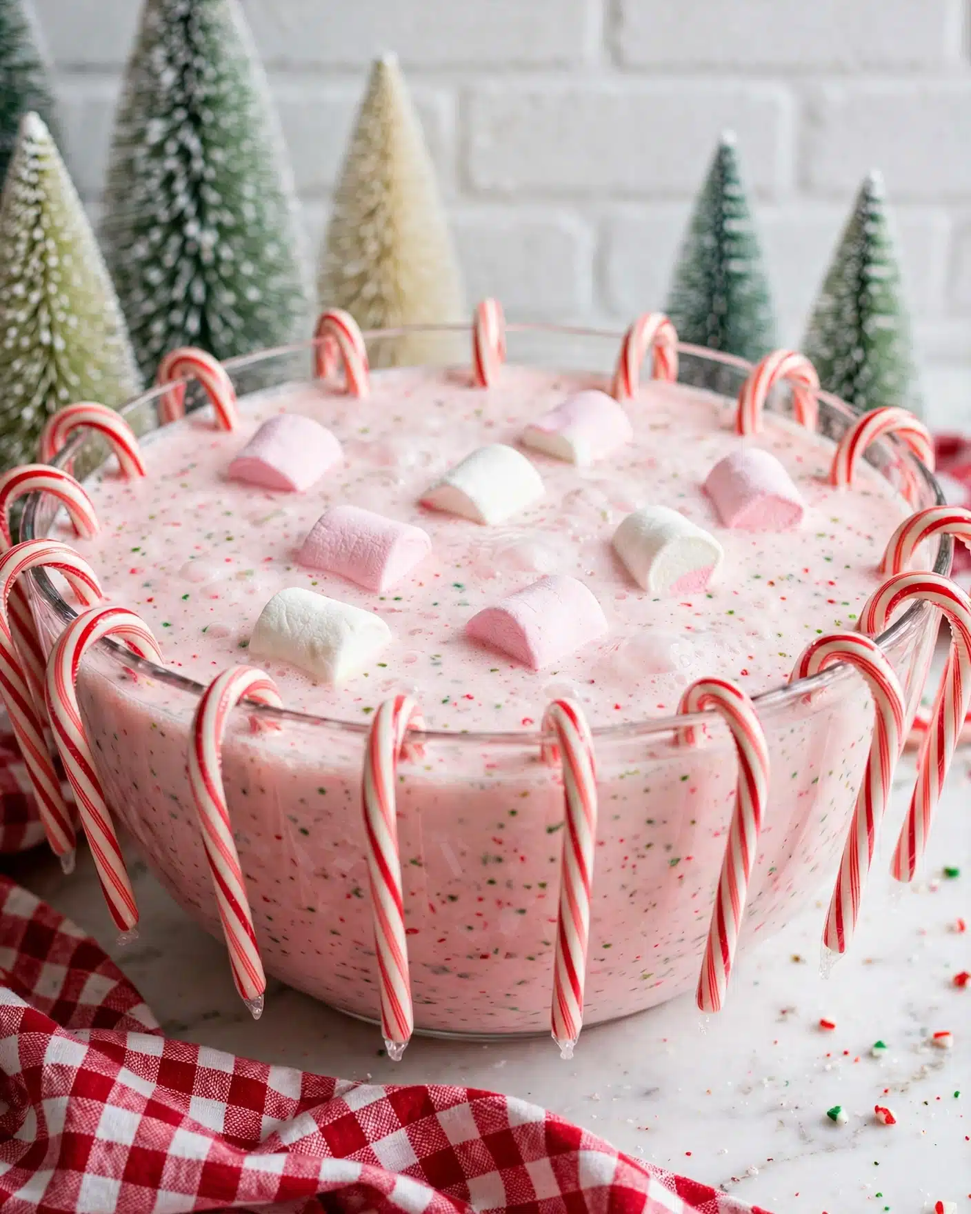 Peppermint Eggnog Punch Recipe 5 A large clear glass bowl filled with light pink foamy punch, topped with some pink and white marshmallows floating on the surface. The bowl is decorated with red and white striped candy canes all around the rim, hanging down its sides. The background shows small green and cream-colored frosted decorative trees against a white brick wall. The bowl sits on a white marbled surface with a red and white checkered cloth placed partially in front. Photo taken with an iphone --ar 4:5 --v 7