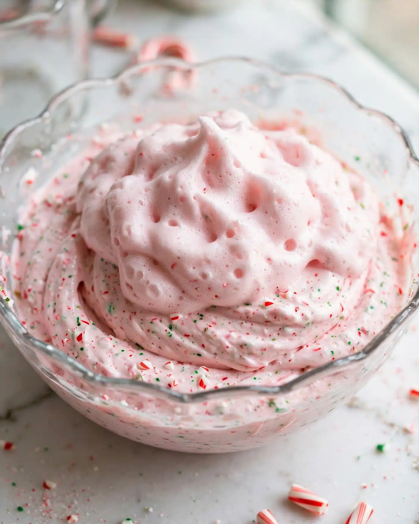 Peppermint Eggnog Punch Recipe 4 A clear glass bowl with a scalloped edge holds a thick, frothy pink mixture with small red and green specks spread throughout. The mixture has a bubbly texture and soft, curved shapes visible under the foam. The bowl is set on a white marbled surface. photo taken with an iphone --ar 4:5 --v 7