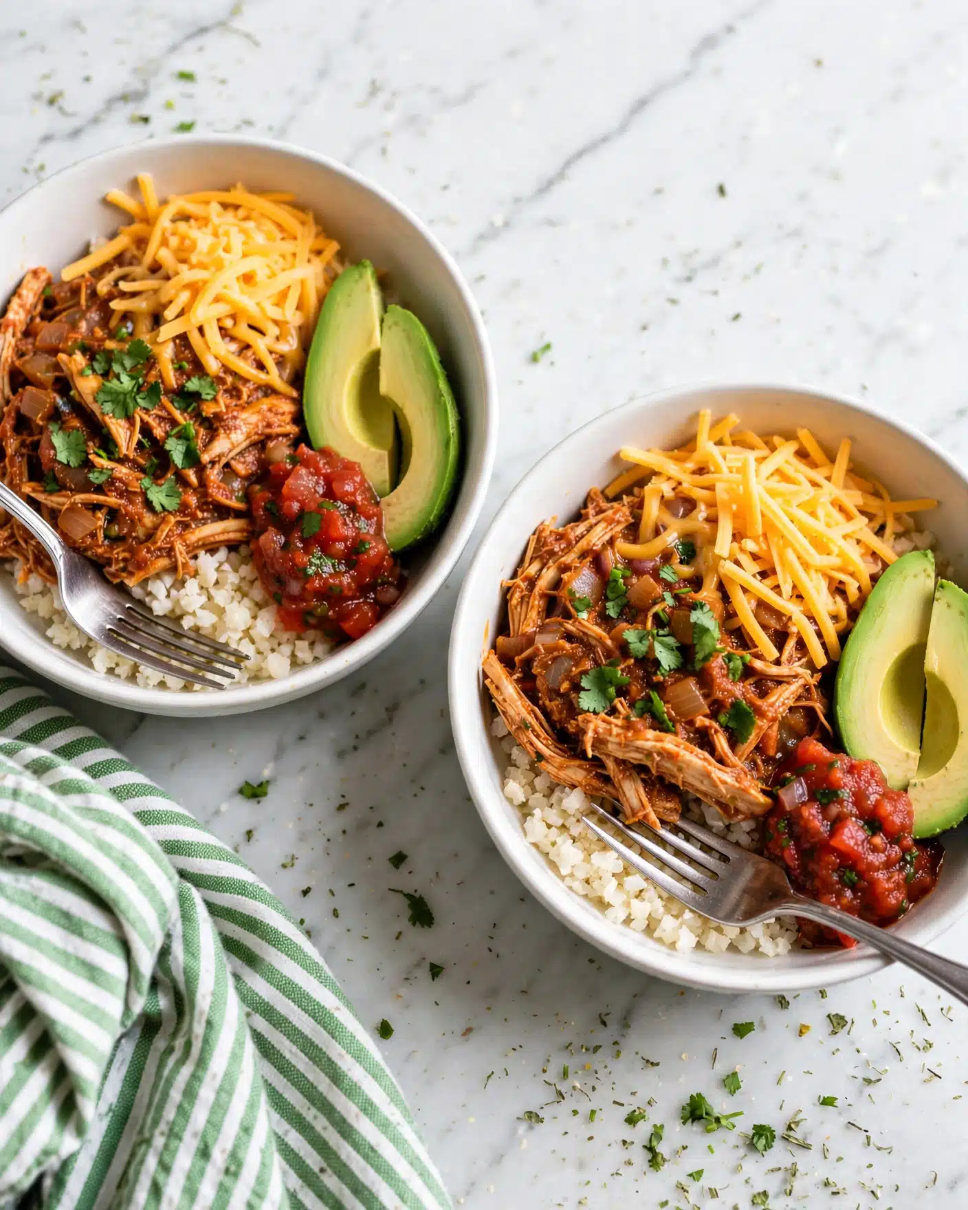 Keto Chicken Enchiladas Bowl Recipe 5 Two white bowls sit on a white marbled surface with green and white striped cloth nearby. Each bowl contains three main layers: at the bottom is a layer of white cauliflower rice, topped with a generous amount of shredded reddish-orange chicken, sprinkled with some fresh green cilantro. On top of the chicken, there is a layer of shredded yellow cheddar cheese. Finally, each bowl has two slices of bright green avocado arranged next to a small dollop of chunky red salsa. Both bowls have a silver fork placed inside. Photo taken with an iphone --ar 4:5 --v 7