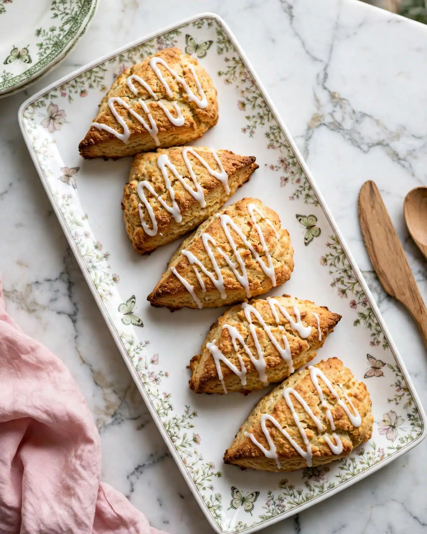 Pumpkin Scones Recipe 5 The image shows five golden-brown scones arranged on a long white plate with floral and butterfly patterns. Each scone is triangular and topped with white icing drizzled in a zigzag pattern, creating a contrast against the textured, slightly cracked surface of the scones. The plate sits on a white marbled surface, and part of another similar white plate with a green patterned edge and a butterfly design is visible in the upper left corner. A pink cloth and light brown wooden utensils are slightly seen on the sides, adding warm tones to the setting. photo taken with an iphone --ar 4:5 --v 7