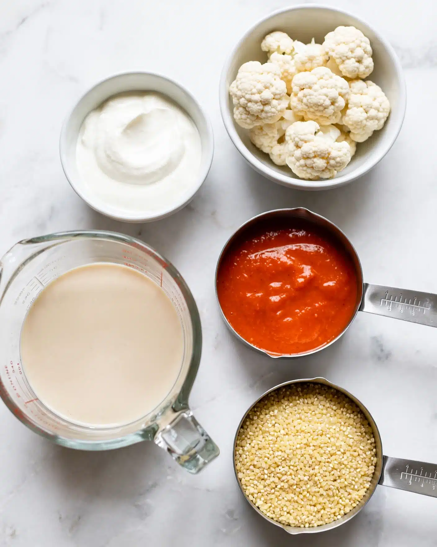 The image shows five white bowls and measuring cups on a white marbled surface, each filled with different ingredients. At the top right, a white bowl is full of small white cauliflower florets. To its left, a smaller white bowl holds a dollop of thick white cream. Below, there is a clear glass measuring cup with a light beige liquid. To the right of the liquid, a metal measuring cup is filled with bright orange-red sauce. Finally, at the bottom right, another metal measuring cup holds small, pale yellow grains, possibly quinoa. All containers are neatly arranged without overlap. Photo taken with an iphone --ar 4:5 --v 7