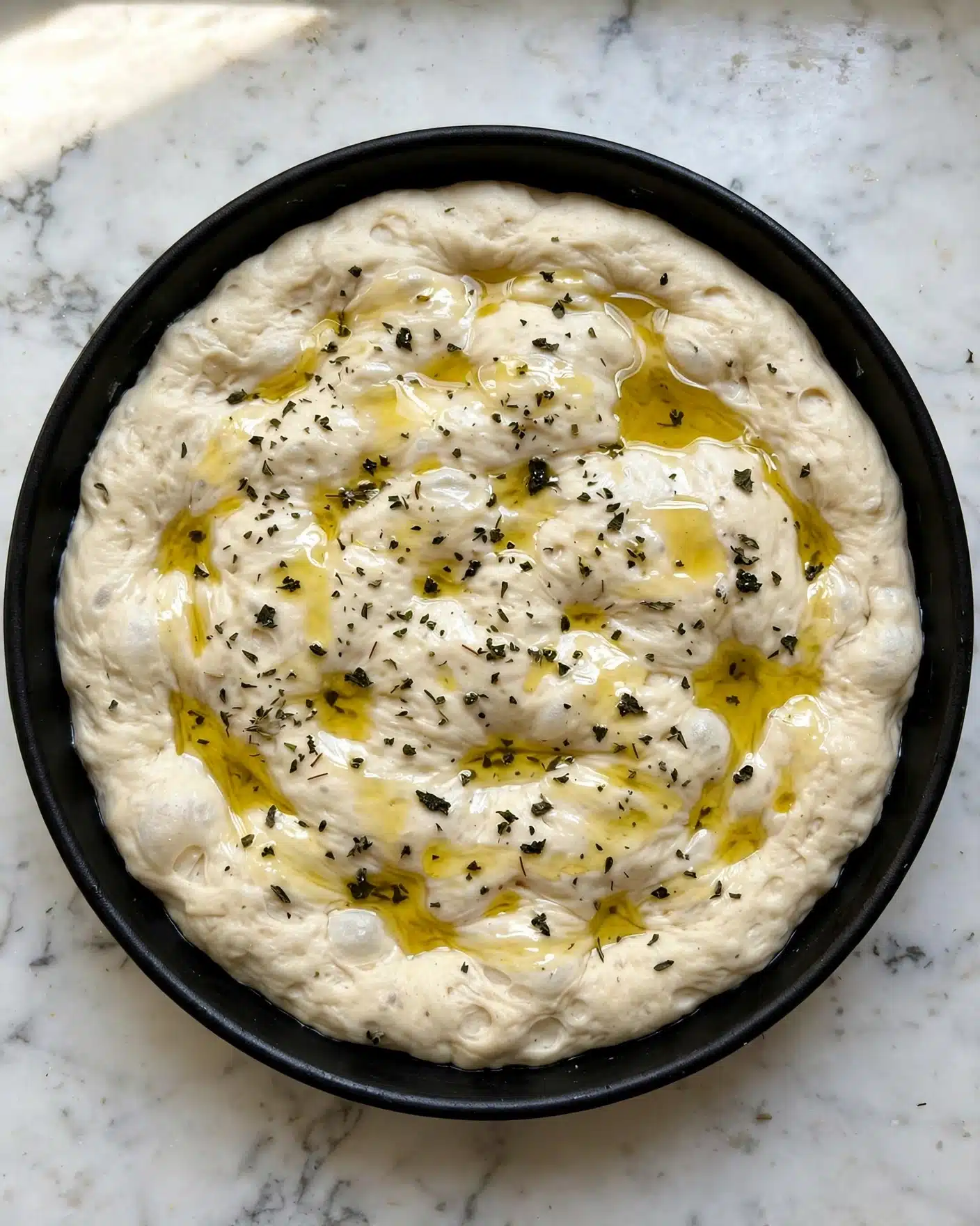 Macaroni and Cheese Pizza Recipe 4 A round black pan holds a single thick layer of pale dough spread evenly inside it, with a smooth and slightly fluffy texture. The dough is sprinkled with small dark green dried herbs and drizzled with golden olive oil in uneven patches, creating a shiny surface with little pools of oil. The pan is placed on a white marbled surface, highlighting the warm earthy tones of the dough and the herbs. photo taken with an iphone --ar 4:5 --v 7