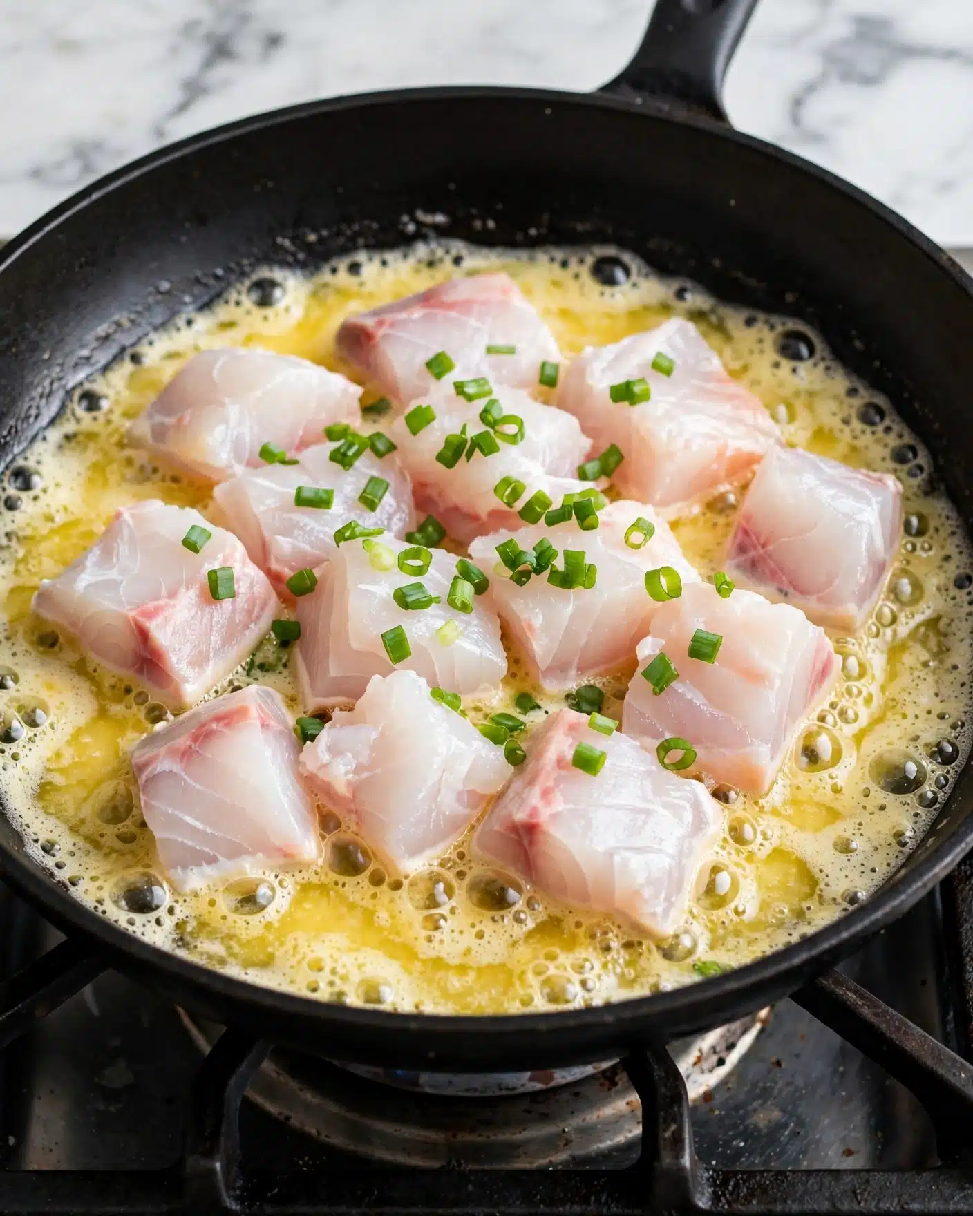 In a black frying pan on a gas stove, there is a cooking mixture with several layers. At the bottom, melted butter is bubbling and has a light golden color. On top of the butter, there are pieces of white raw fish, some with a slight pink tint, and a few green chopped scallions scattered over the fish. The texture of the fish looks soft and fresh, while the scallions add a bright contrast. The background is a white marbled texture. Photo taken with an iphone --ar 4:5 --v 7