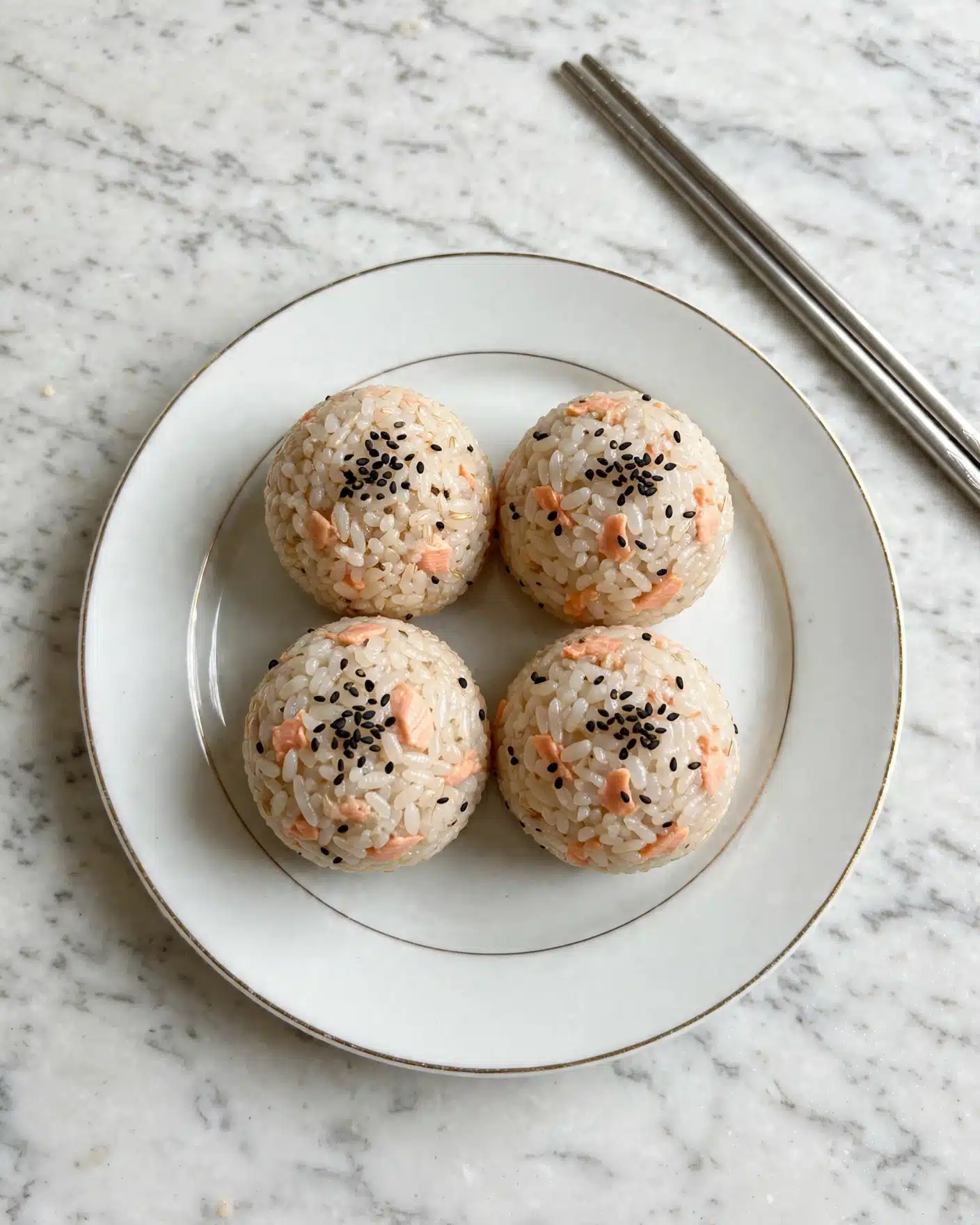Salmon Rice Balls Recipe 5 There are four round rice balls on a white plate with a thin gold edge, each ball made from light brown rice mixed with small pieces of light pink salmon, and topped with a small sprinkle of black sesame seeds; the plate is placed on a white marbled surface, with a pair of silver chopsticks visible in the upper right corner photo taken with an iphone --ar 4:5 --v 7