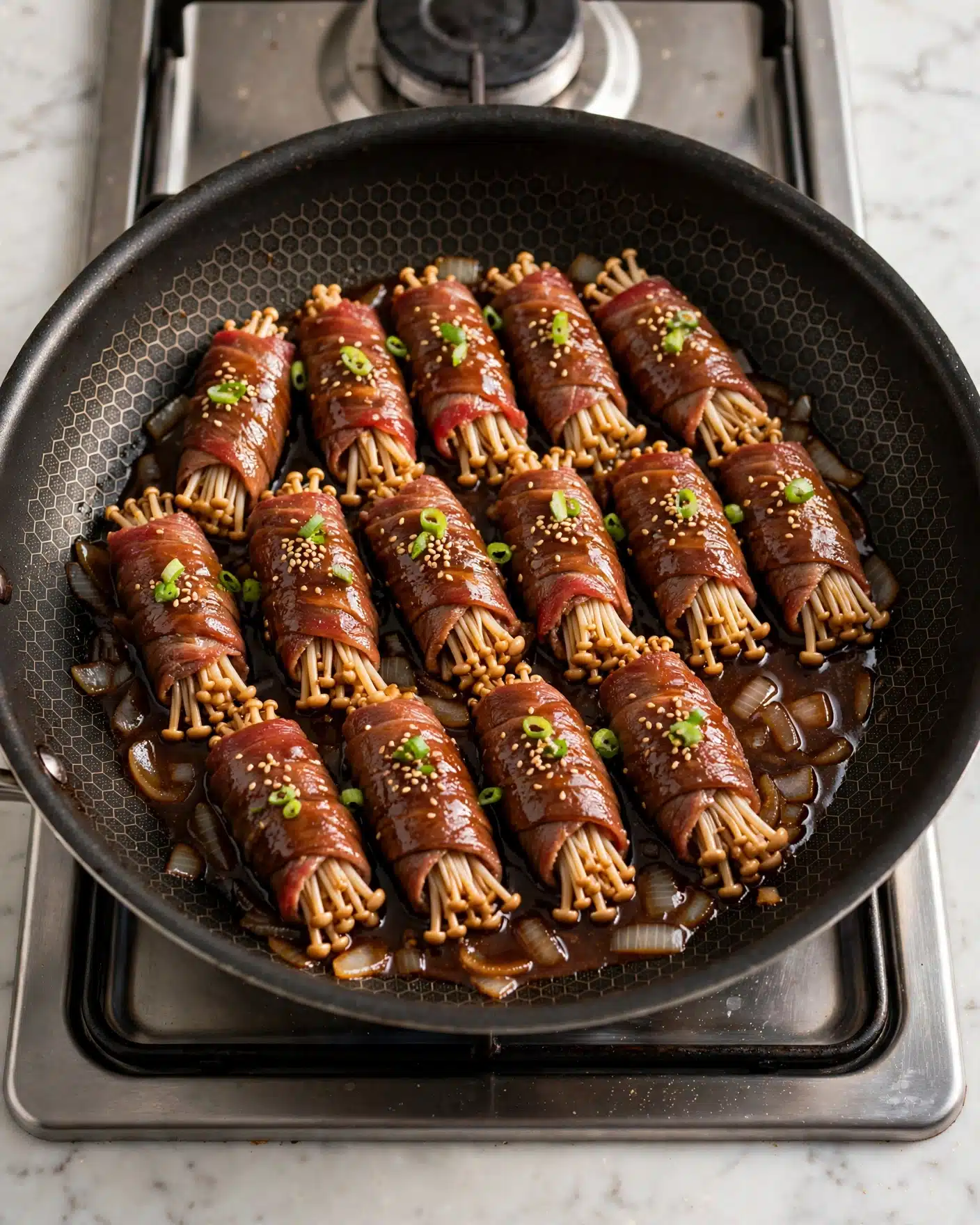 Enoki Beef Rolls in Dashi Soy Sauce Recipe 4 In a black frying pan with a honeycomb pattern, there are fifteen rolls made by wrapping thin red meat around bundles of small white mushrooms. The rolls are arranged in three staggered rows, each resting in a dark brown sauce with small pieces of translucent cooked onions scattered underneath. The pan is placed on a silver stove burner, and the whole scene is on a white marbled surface. photo taken with an iphone --ar 4:5 --v 7