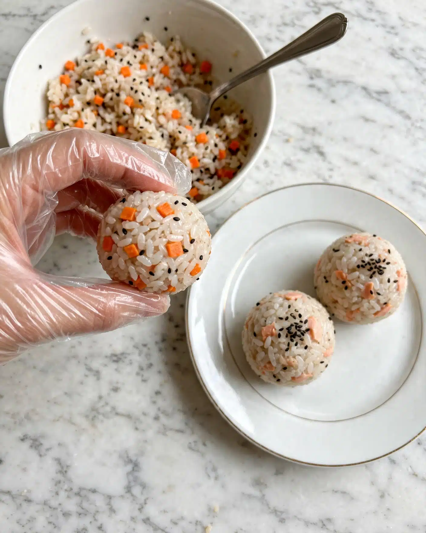 Salmon Rice Balls Recipe 4 A woman's hand wearing a clear plastic glove holds a small round ball made of white rice mixed with small orange carrot pieces and a few black sesame seeds. In the background, there is a white bowl filled halfway with more of the same rice mixture and a silver spoon inside it. To the right, on a white plate, there are two more rice balls placed next to each other. The whole scene is set on a white marbled surface. photo taken with an iphone --ar 4:5 --v 7