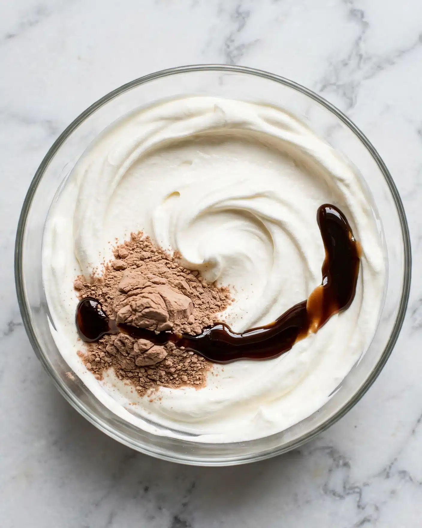 Chai Latte Cupcakes Recipe 4 A clear glass bowl holds three separate layers of ingredients on a white marbled surface: a large swirl of smooth, white cream covering most of the bowl's bottom, a mound of light brown powder positioned near the lower left side of the cream, and a glossy dark brown liquid poured in a curved line crossing over part of the powder and cream near the right center. The textures contrast between the thick cream, fine powder, and shiny liquid, all waiting to be mixed together. Photo taken with an iphone --ar 4:5 --v 7