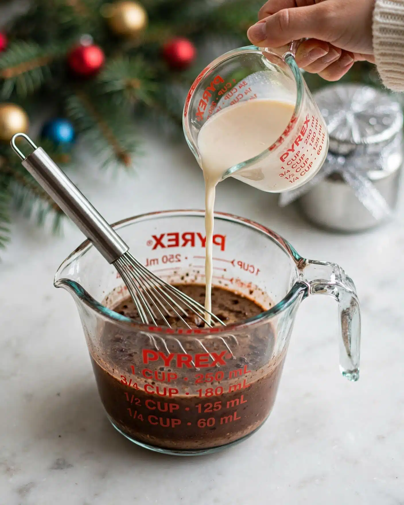 The image shows a transparent glass Pyrex measuring cup with red measurement markings. Inside the cup, there is a dark brown liquid mixture being stirred with a metal whisk. A woman's hand is pouring a light cream-colored liquid from another glass measuring cup into the cup with the brown liquid, creating a small creamy stream. The scene is set on a white marbled surface with soft natural light. photo taken with an iphone --ar 4:5 --v 7