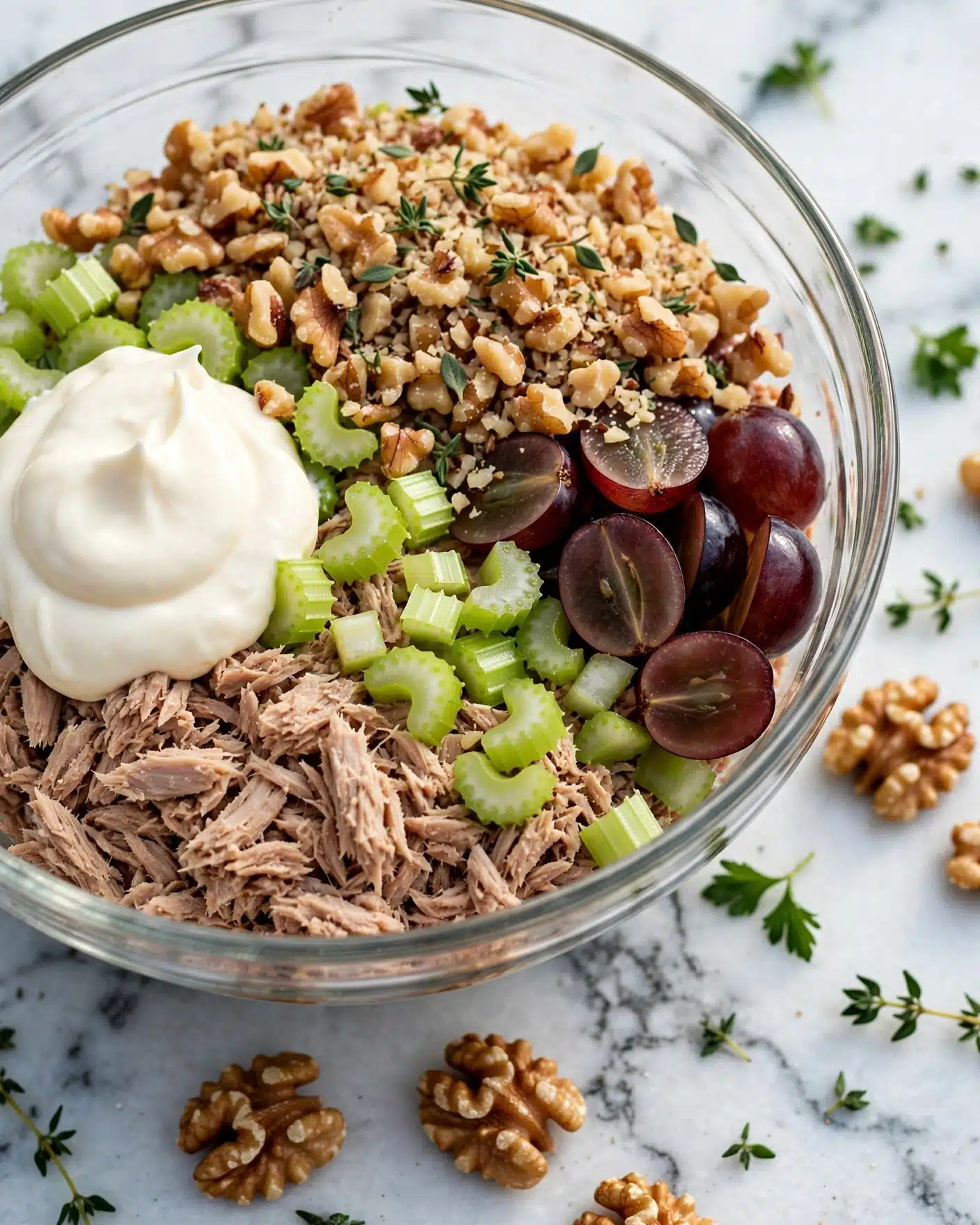 California Chicken Salad Recipe 4 A clear glass bowl sits on a white marbled surface, showing a mix of ingredients inside. The bottom layer is shredded light brown tuna, topped on one side with a dollop of smooth white mayonnaise. Next to it, there are small bright green celery pieces scattered, and on the other side, halved dark red grapes with shiny skins. The last top layer is chopped light brown walnuts spread over half the bowl, sprinkled with tiny green thyme leaves. A few whole walnut pieces and a thyme sprig lay on the surface outside the bowl. photo taken with an iphone --ar 4:5 --v 7