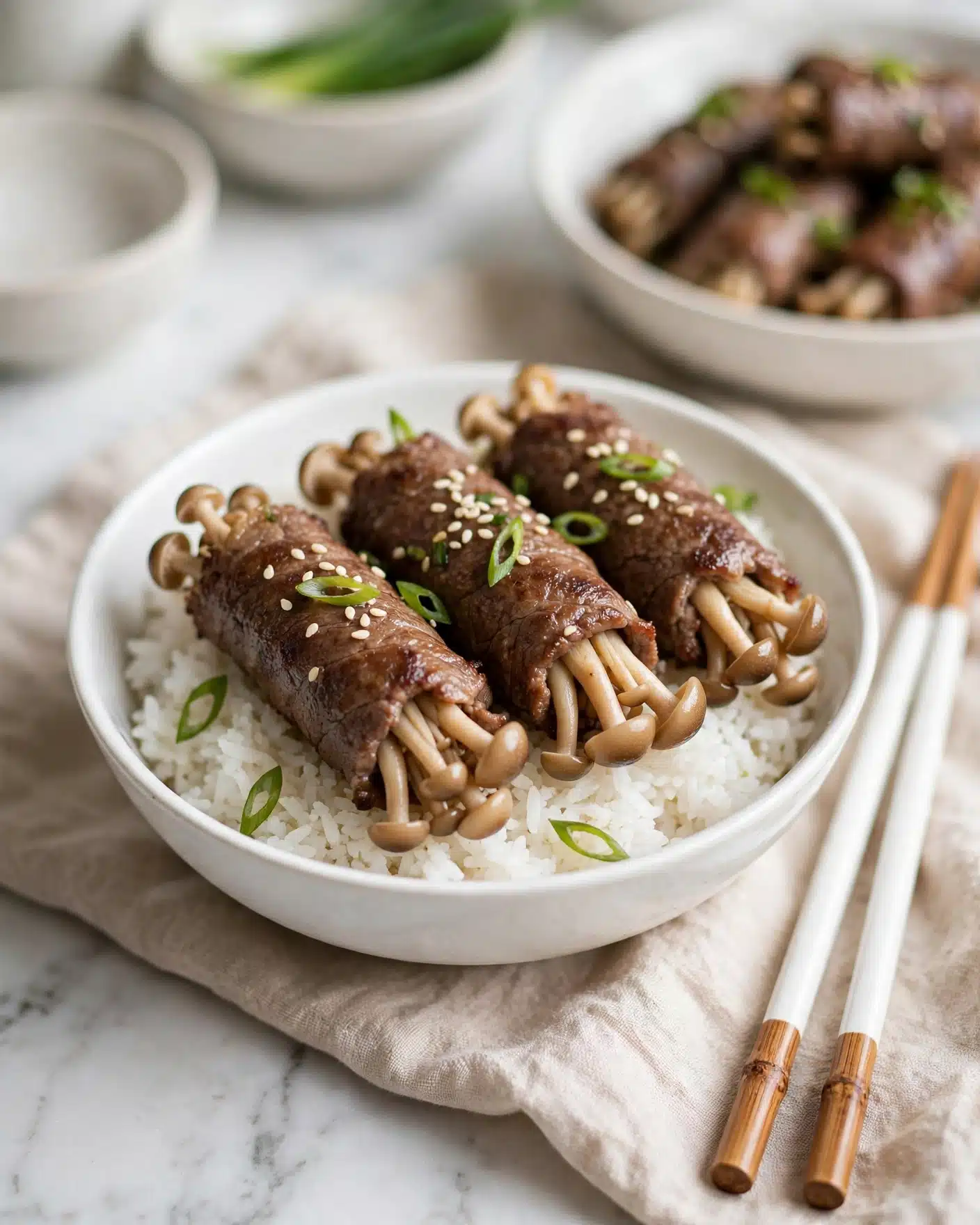 Enoki Beef Rolls in Dashi Soy Sauce Recipe 5 A white bowl filled with a base layer of fluffy white rice, topped with four brown beef rolls wrapped around clusters of light tan mushrooms. The rolls are garnished with small white sesame seeds and thin green slices of scallion scattered across the top. The bowl is placed on a soft beige cloth on a white marbled surface, with a pair of white chopsticks with wooden tips resting nearby. In the background, there are blurred white bowls containing green scallions and more of the beef roll dish. Photo taken with an iphone --ar 4:5 --v 7