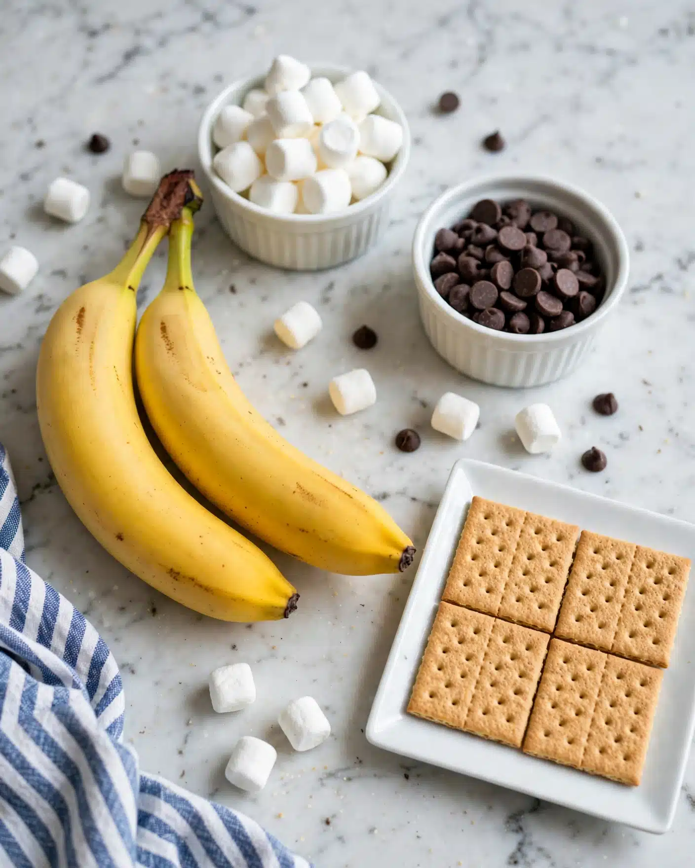 The image shows four light brown graham crackers laid flat on a white rectangular plate at the bottom right. To the left of the plate, there are two bright yellow bananas with green stems lying side by side on a white marbled surface. Above the bananas, there is a small white bowl filled with small white marshmallows, and to its right, another small white bowl filled with dark brown chocolate chips. A blue and white striped cloth is partially visible in the bottom left corner. The whole scene is set on a white marbled textured background. photo taken with an iphone --ar 4:5 --v 7