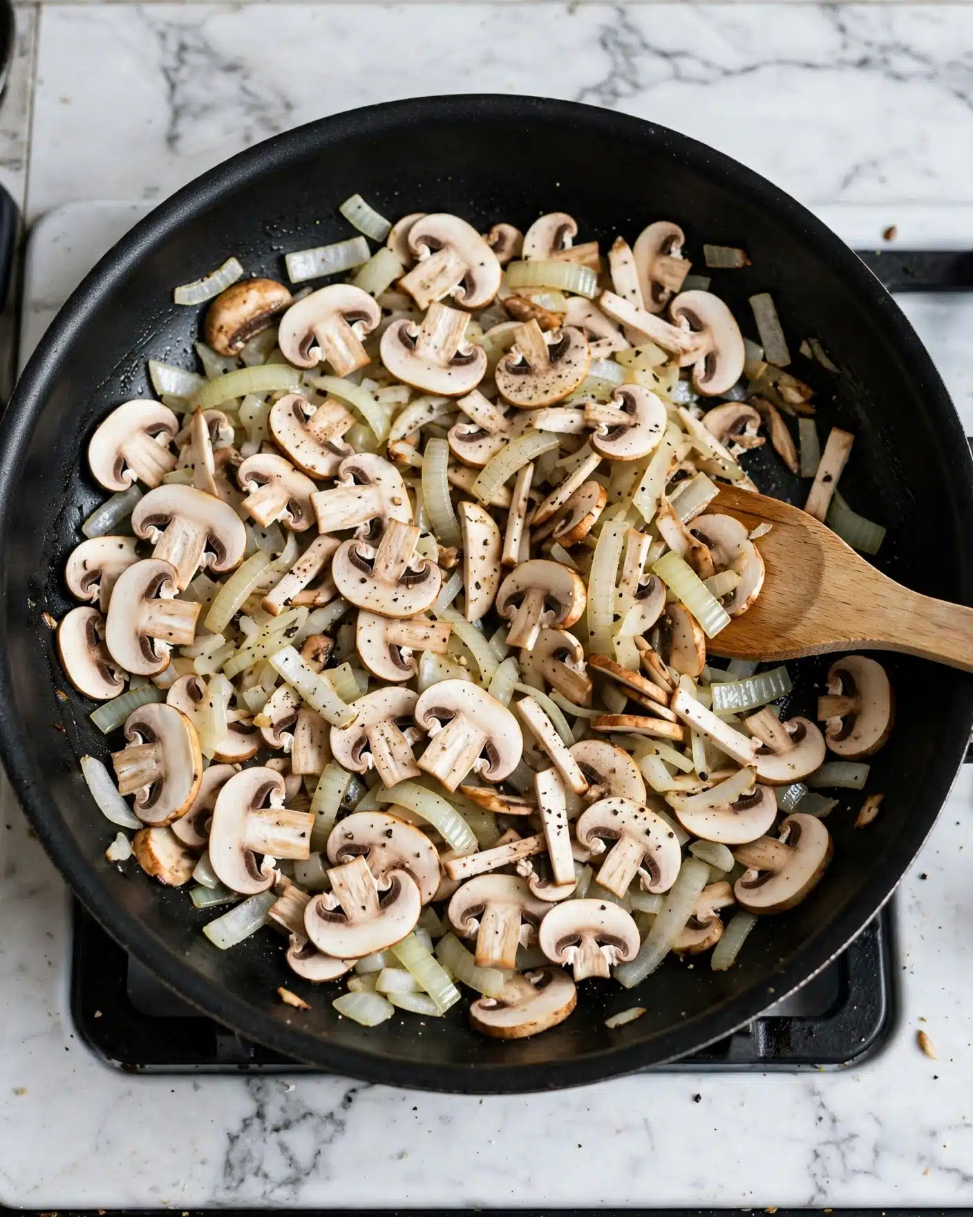 A black pan placed on a stove filled with many sliced white mushrooms mixed with some pieces of onion. The mushrooms are spread out evenly in the pan, showing a fresh white and light brown color. There is a light sprinkling of black pepper on top of the mushrooms. A wooden spatula is partially inside the pan on the right side, touching the mushroom layer. The stove surface around the pan is white marbled texture photo taken with an iphone --ar 4:5 --v 7
